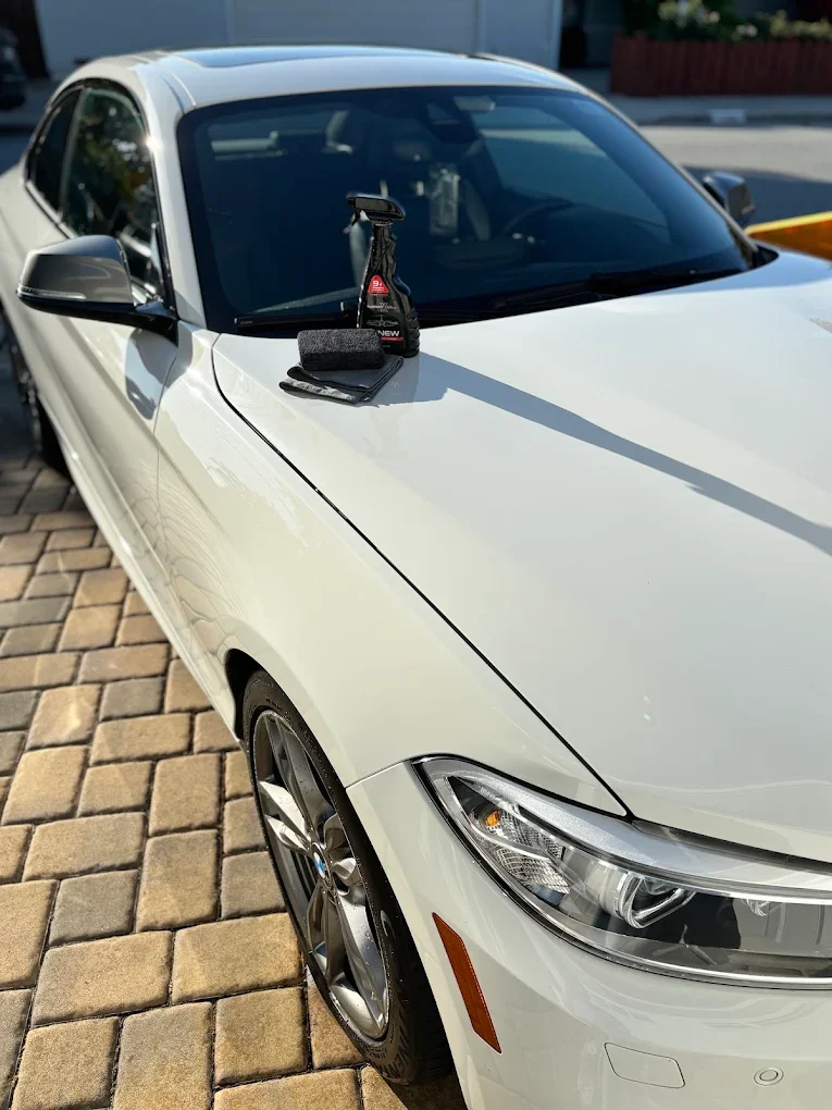 White sedan car parked on brick pavement with cleaning supplies on hood, including spray bottle, cloth, and cleaning tool.