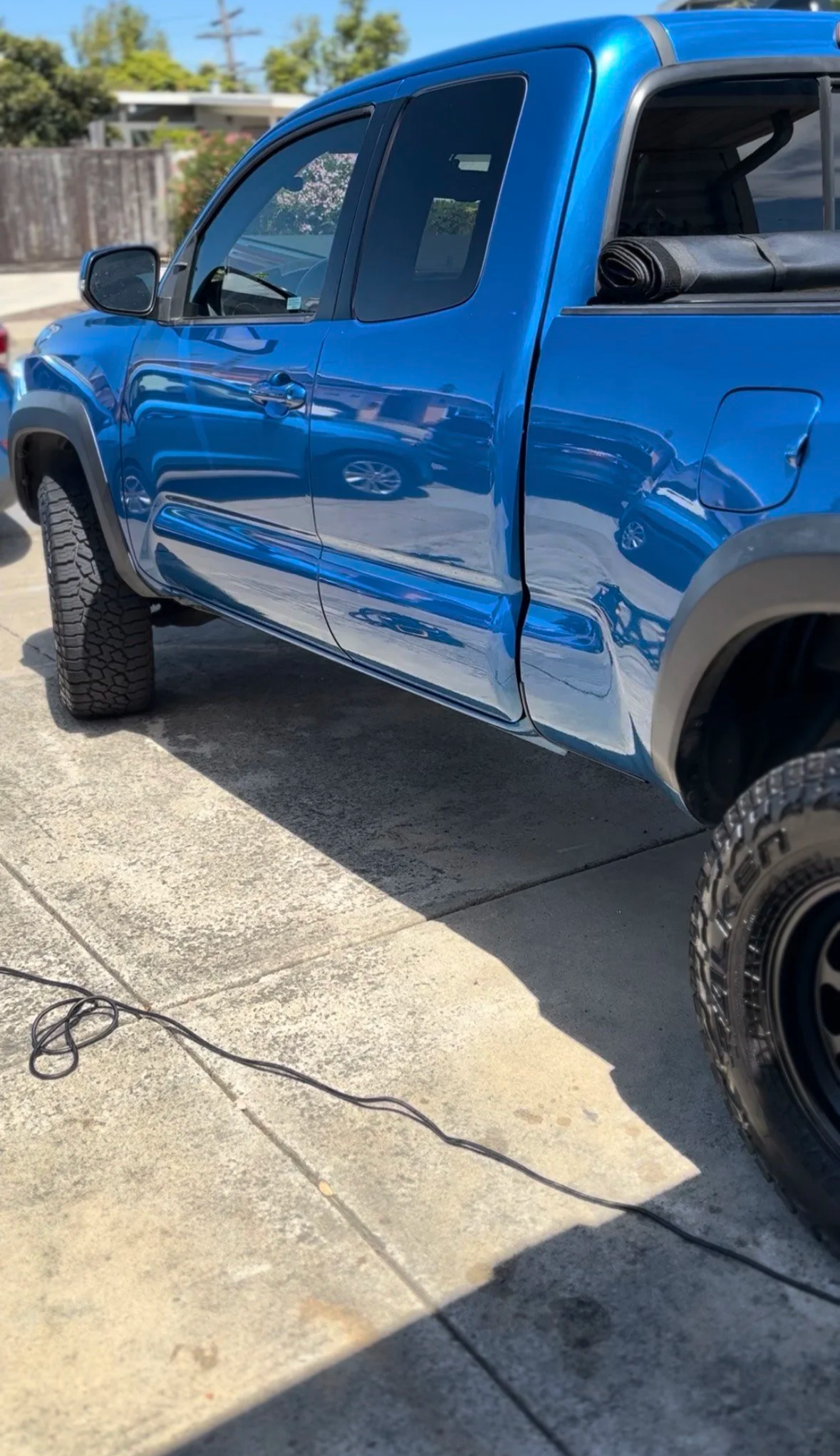 Blue pickup truck parked on a concrete driveway, with a rolled-up mat or tent stored in the truck bed, and a black power cord lying on the ground nearby.