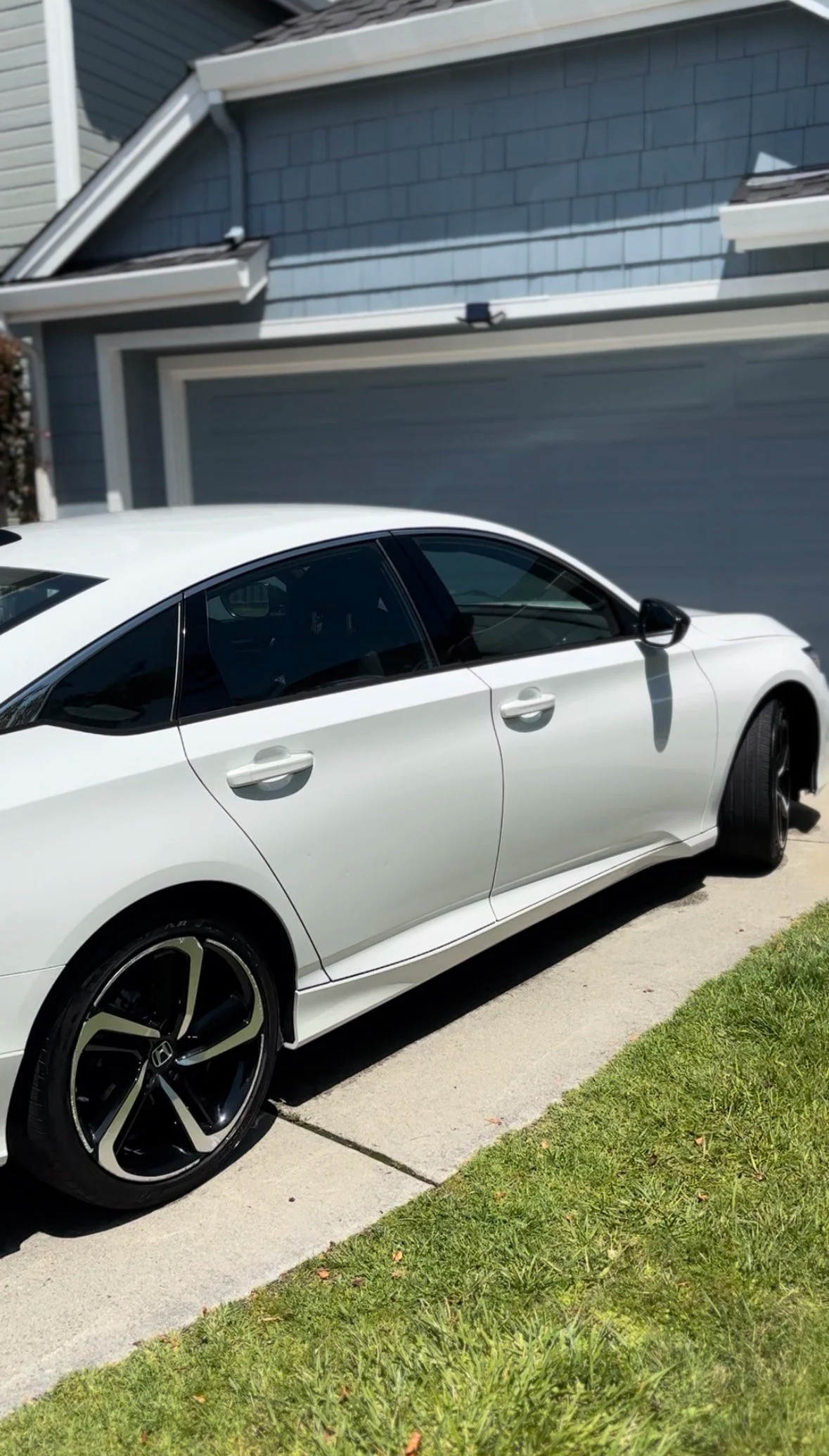 A white Honda sedan parked in a driveway next to a grass lawn, with a blue garage door and house siding in the background.
