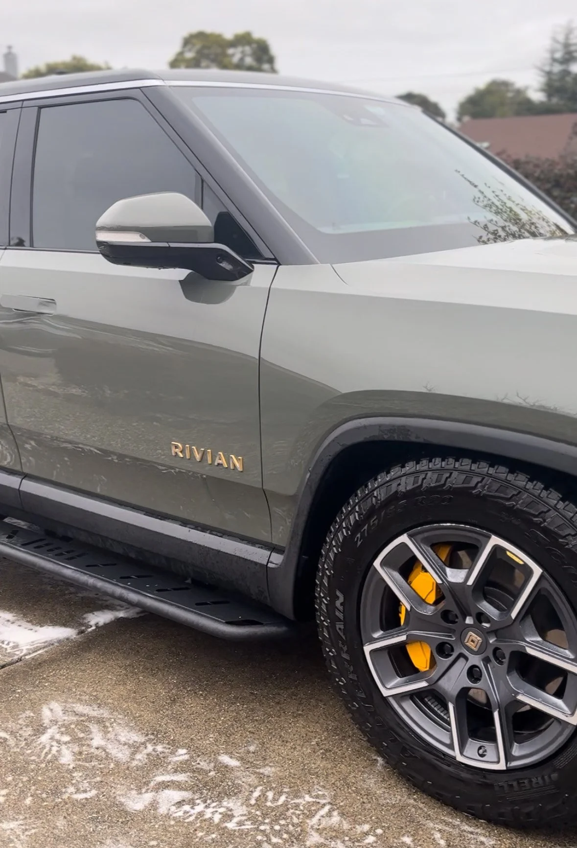 Close-up of a gray Rivian electric vehicle, showing the front side, wheel, and side mirror, parked on a wet street with partial snow.