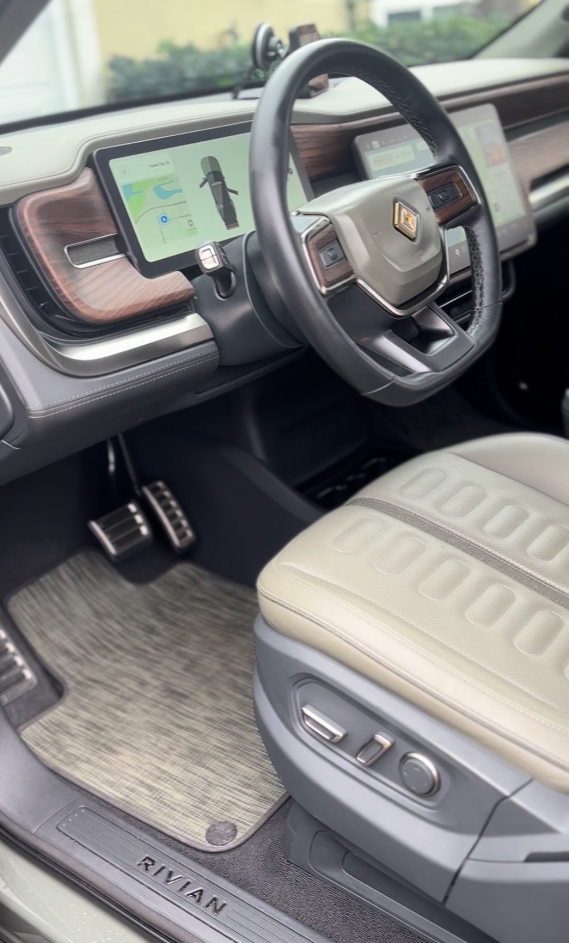 Interior of an electric vehicle with a steering wheel, digital dashboard display, beige seat with control buttons, and RIVIAN branding on the door sill.