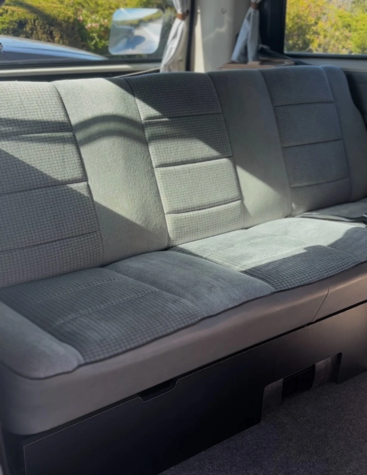 Interior of a vehicle showing a bench seat with a patterned fabric upholstery and a window with a view of green bushes outside.