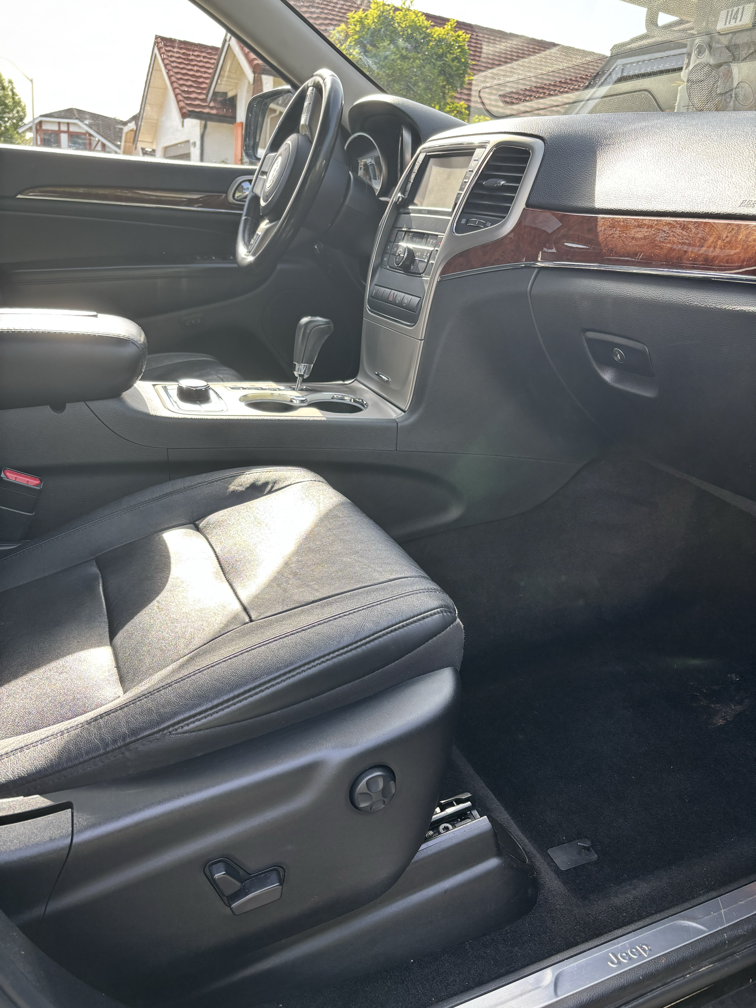 Interior of a Jeep vehicle showing the driver's seat, dashboard, steering wheel, center console with gear shift, and part of the outside neighborhood through the windshield.