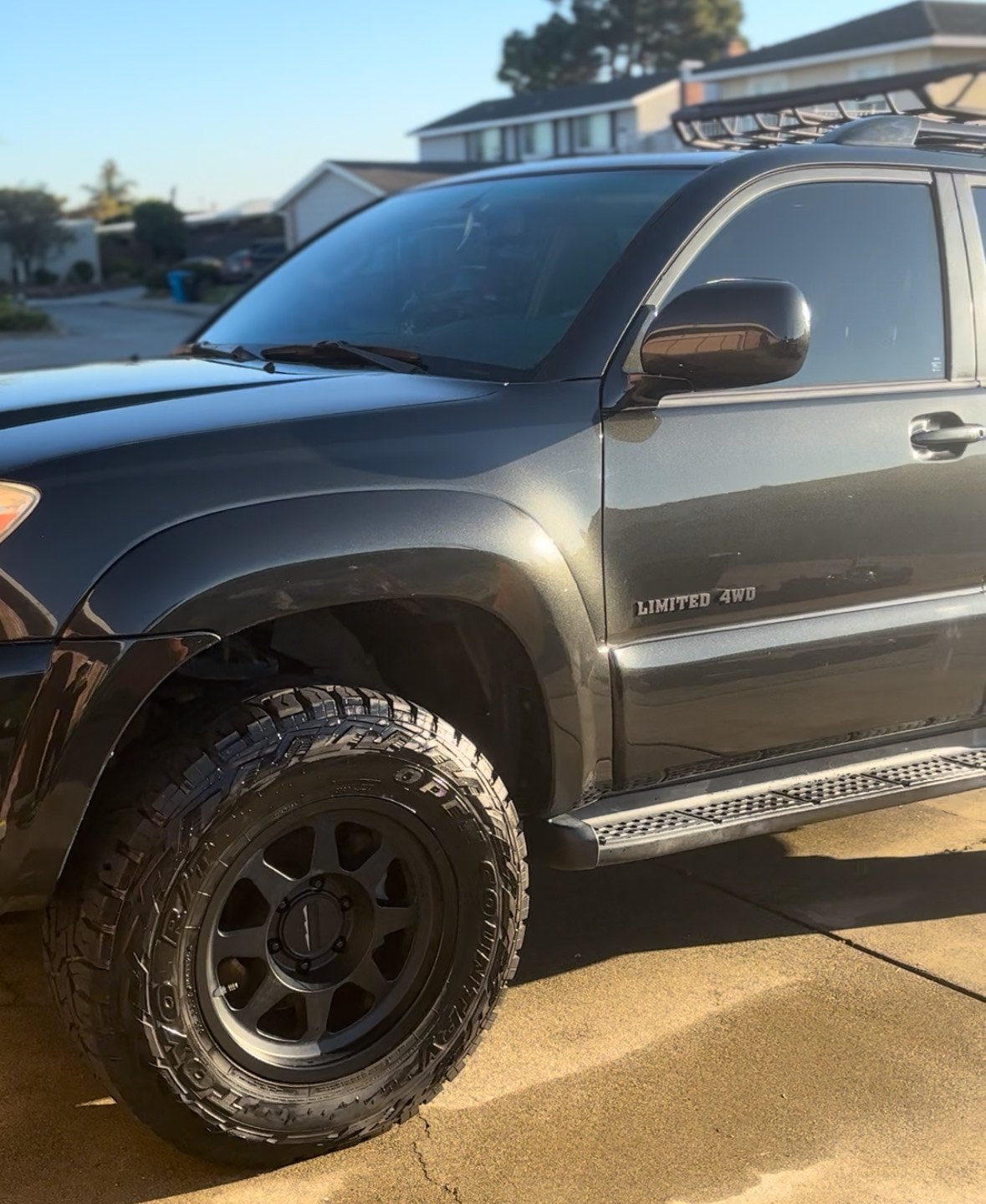 Close-up of a black SUV with large off-road tires, parked on a driveway in front of a suburban house.