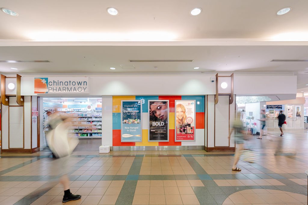 Entrance to Chinatown Pharmacy inside a shopping mall, with posters on the wall and several blurred people walking.