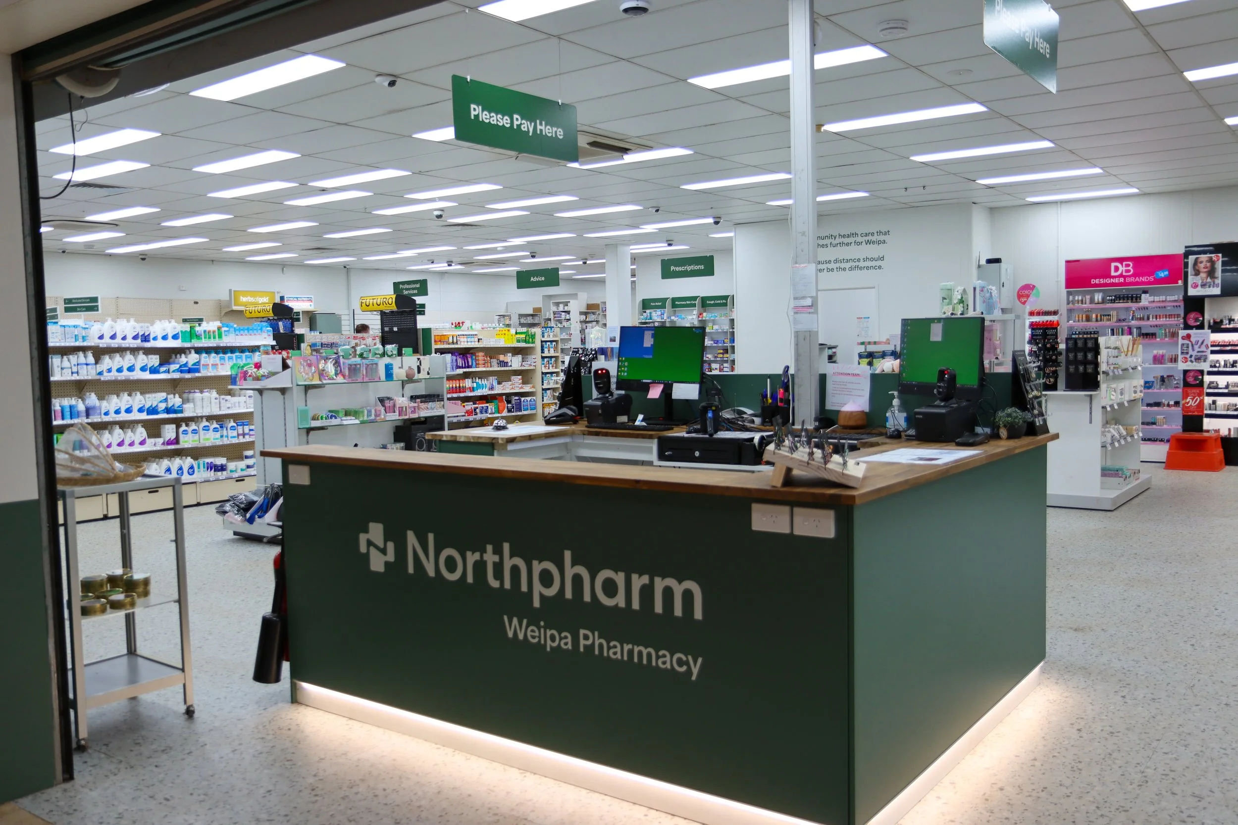 Inside a pharmacy, storefront counter with computer monitors, signs for prescriptions and advice, shelves with various health products, and a green sign overhead that says 'Please Pay Here'.