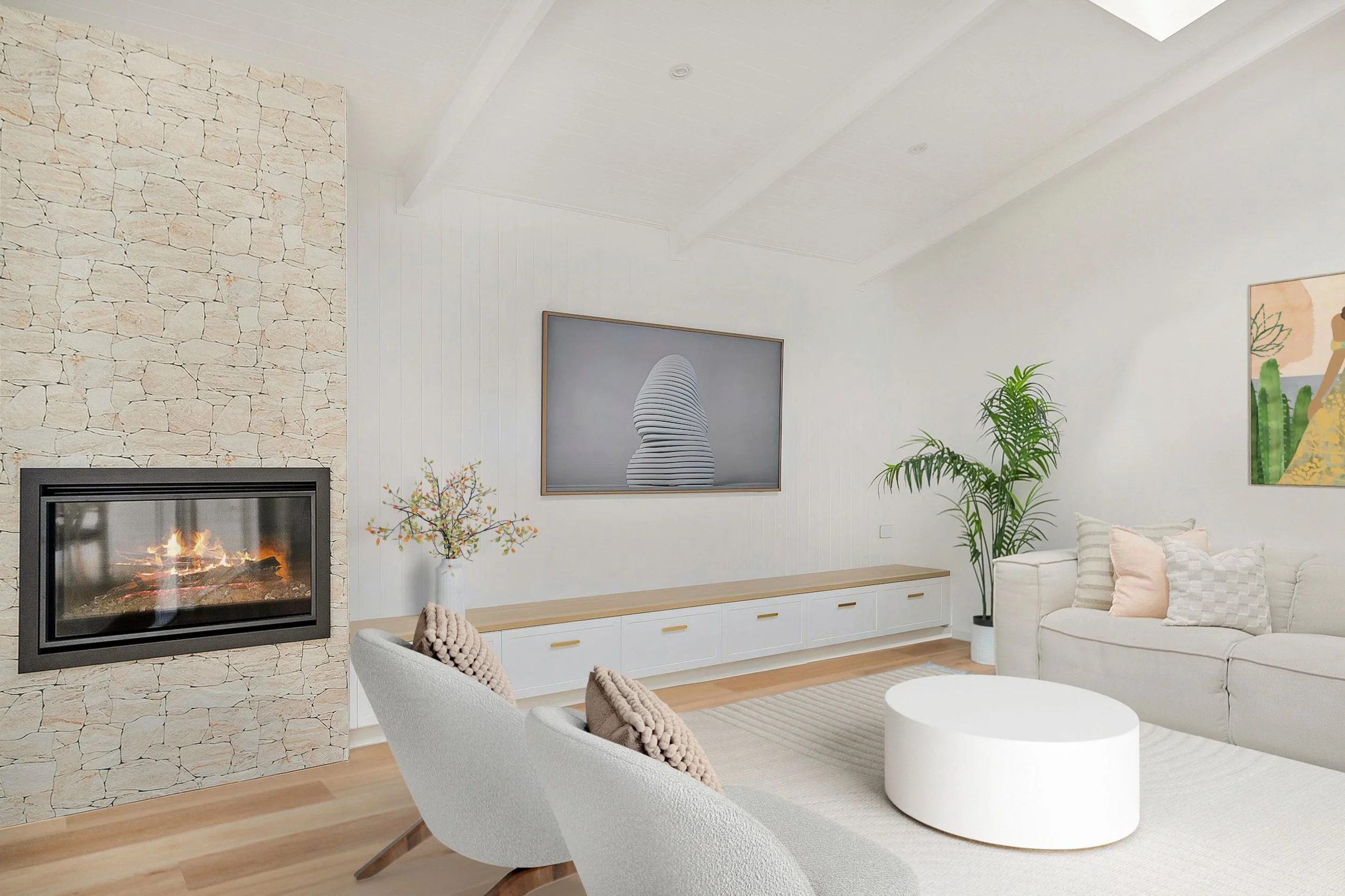 Living room with a stone fireplace, white walls, a beige couch with pillows, two white armchairs, a large insert TV, artwork, plants, and a low round white coffee table.