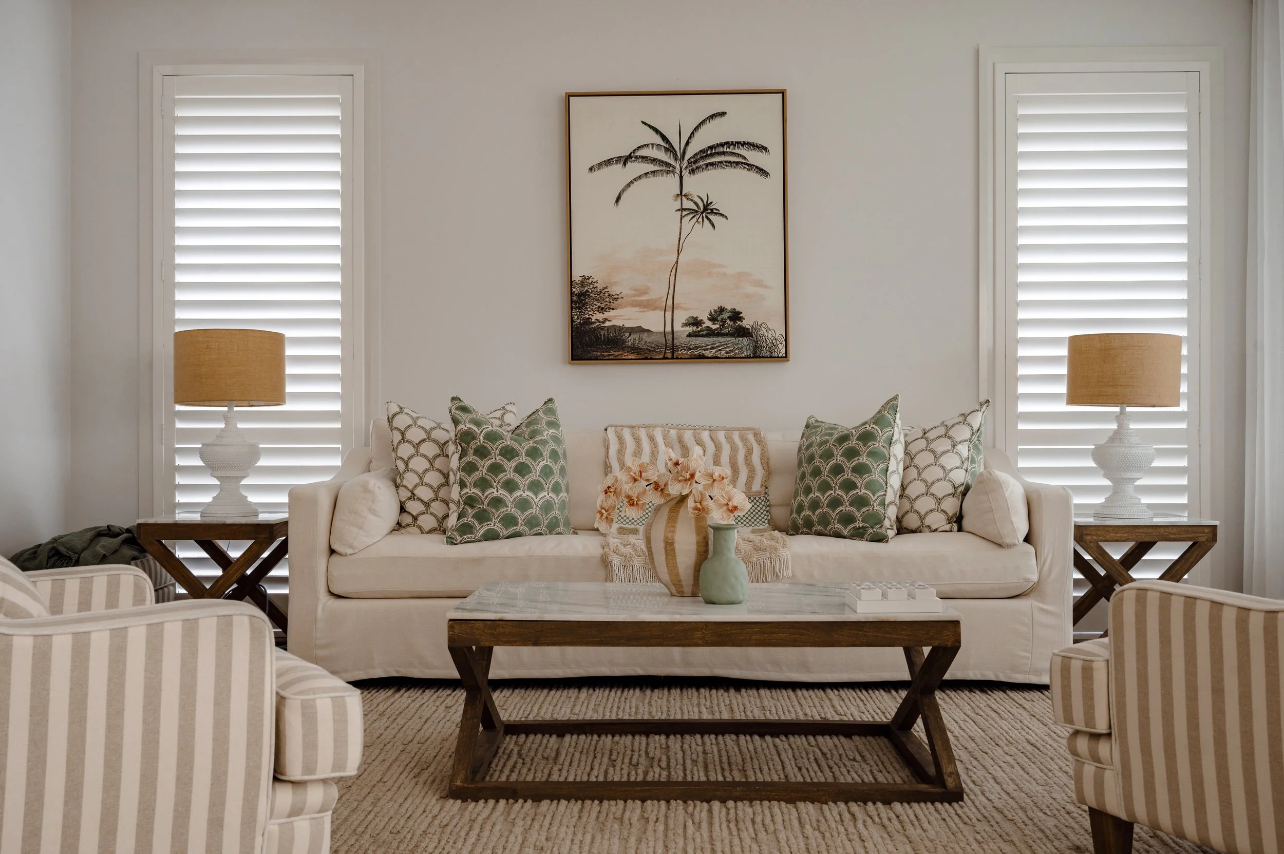 Living room with beige sofa and striped chairs, green patterned pillows, wooden coffee table, table lamps, framed palm tree artwork, window shutters, and beige rug.