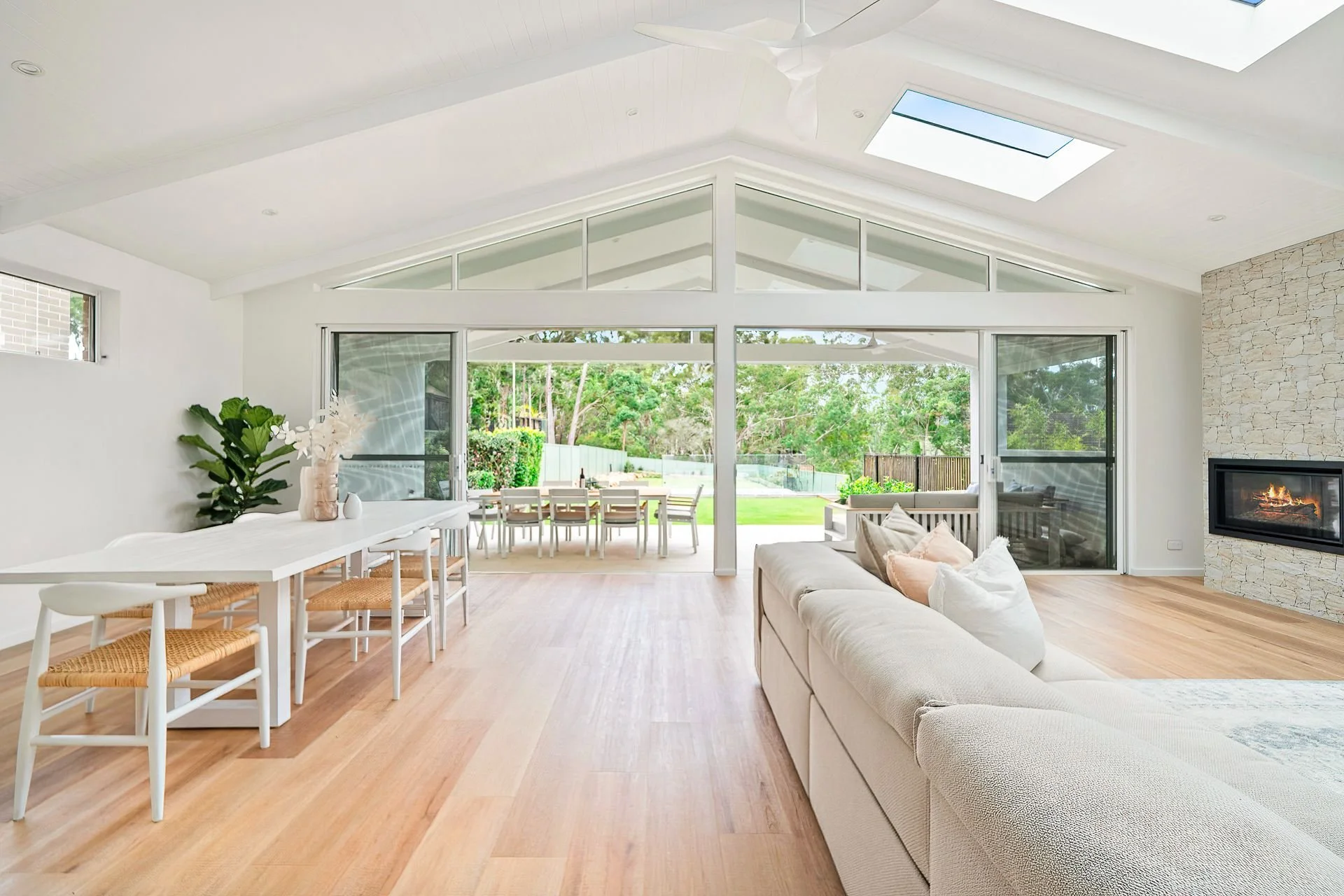 Bright living room with white walls, wooden flooring, a white sofa with pillows, a stone fireplace, large glass doors opening to a backyard patio with outdoor dining area and trees.