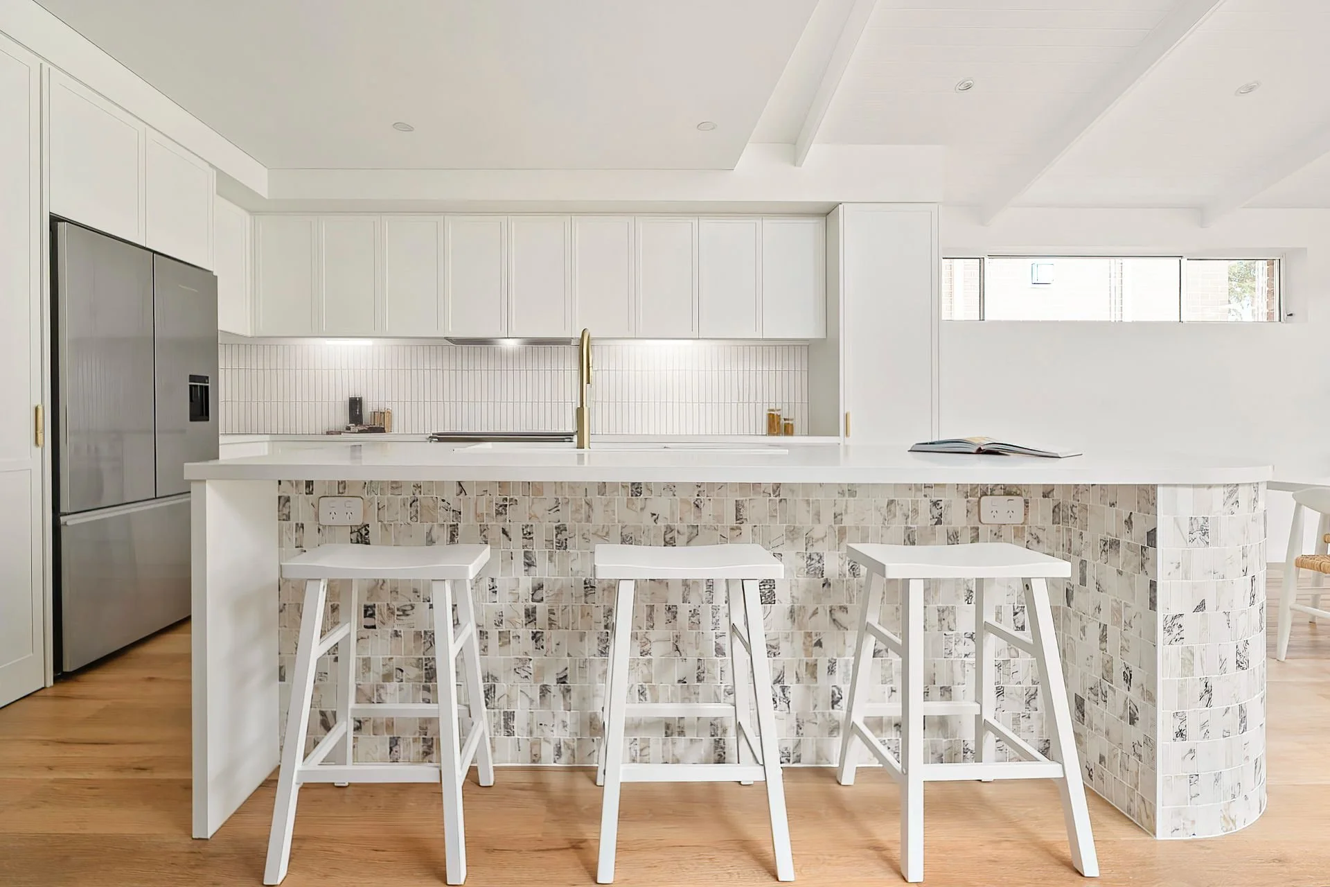 Modern white kitchen with a large island, three white bar stools, a white backsplash, and a stainless steel refrigerator.