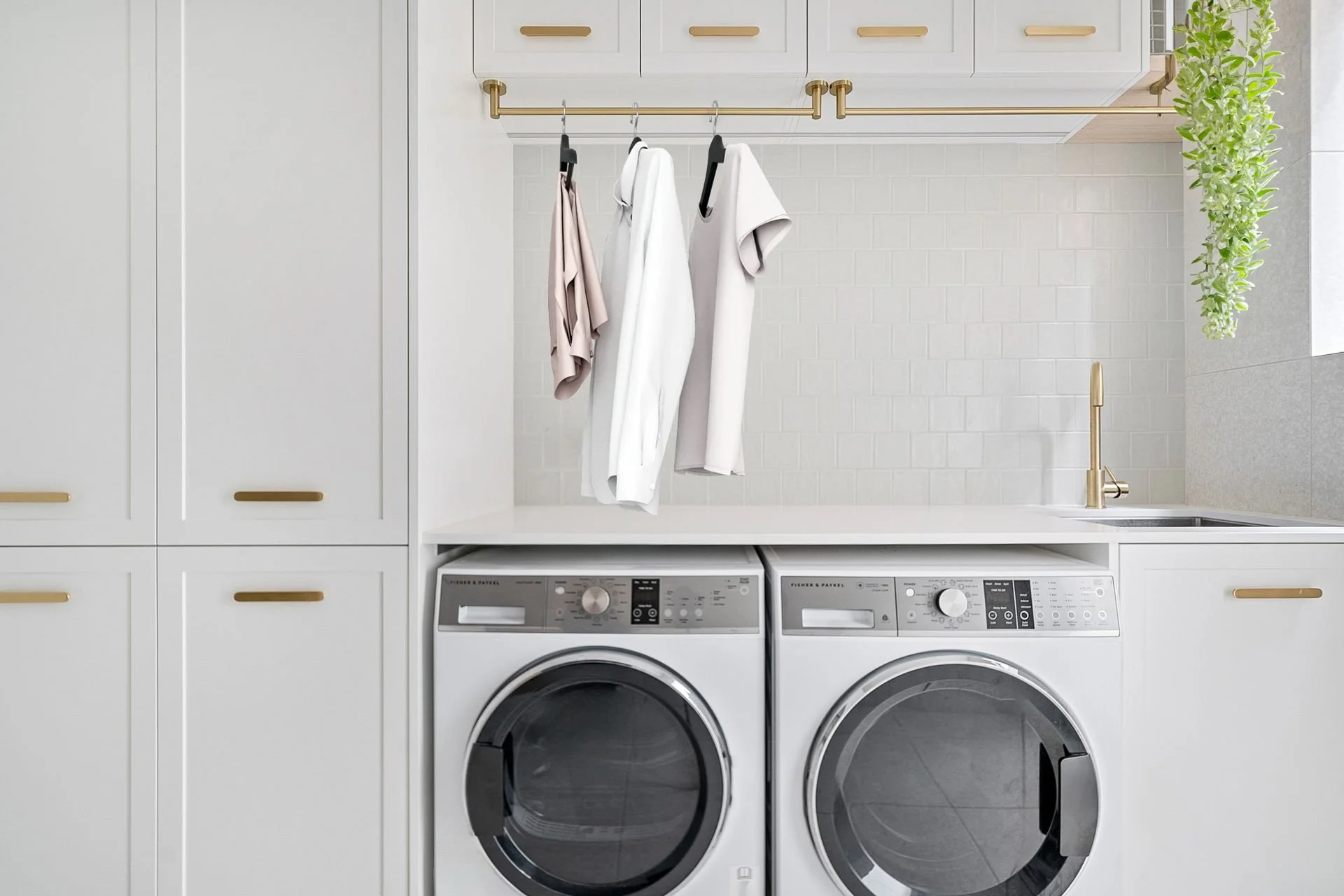 A modern laundry room with white cabinets, a gold hanging rod with clothes, a gold faucet at a sink, and a plant on the side. Two front-loading laundry machines are beneath the white countertop.