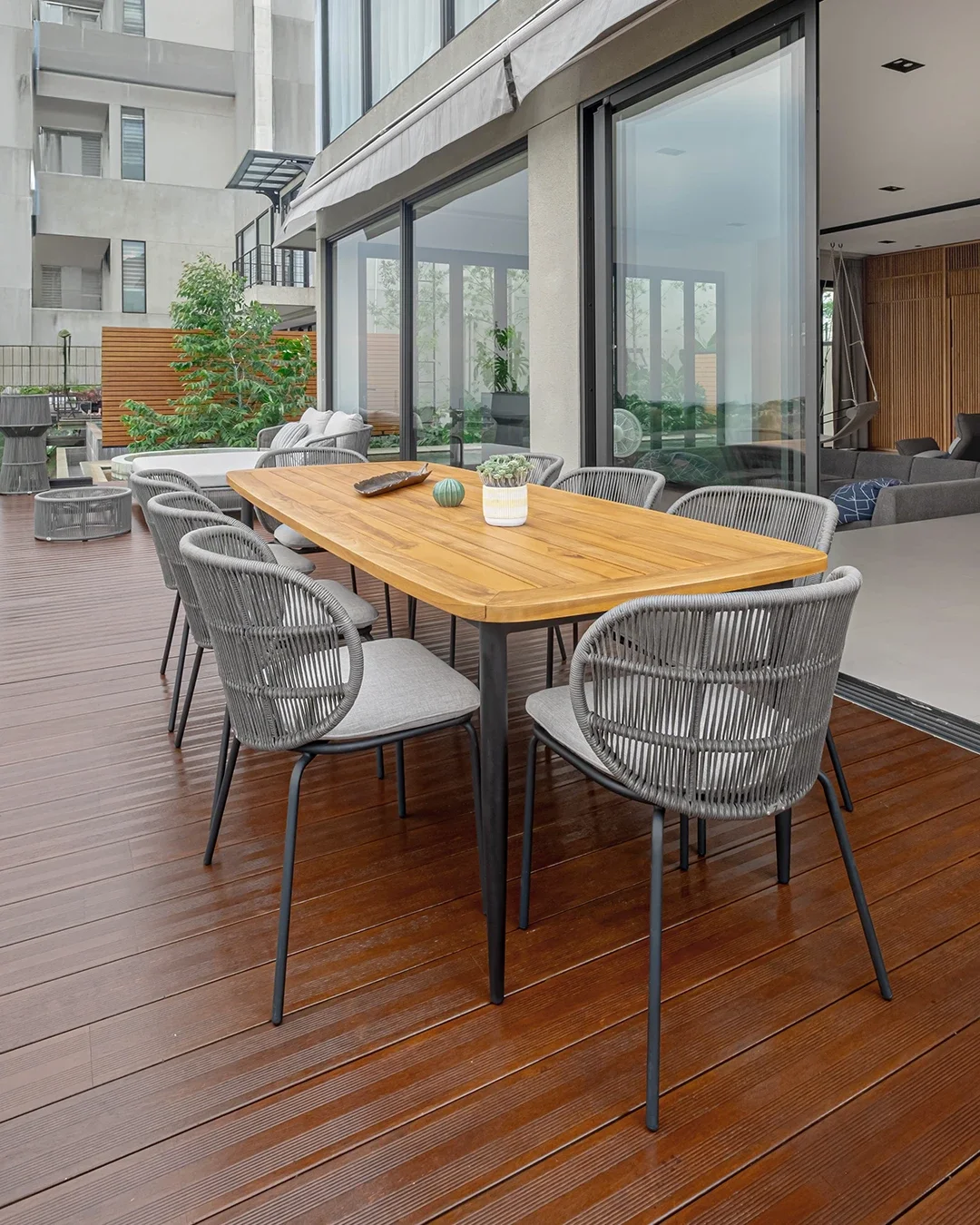 Outdoor patio with a wooden dining table, eight gray woven chairs, potted plants, and a seating area with lounge chairs and a hammock, adjacent to a modern apartment building.