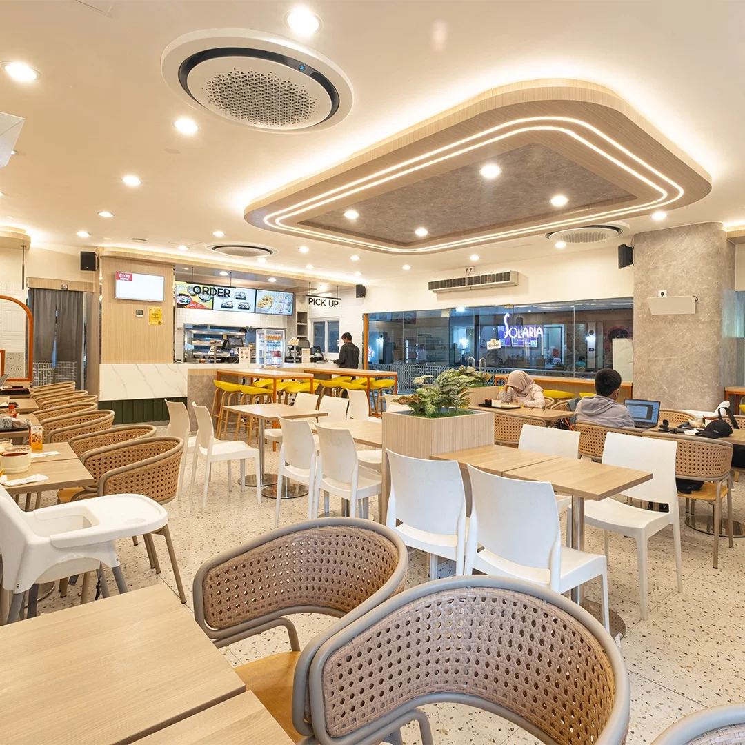 Inside a modern, well-lit restaurant or cafe with multiple tables and chairs, a counter at the back for ordering and pickup, and a few customers seated and working on laptops.