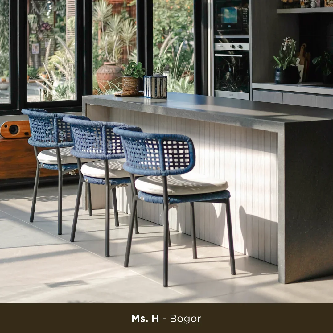 Modern kitchen with a light gray counter island, three blue woven bar stools with white cushions, potted plants, and large windows showing a garden outside.