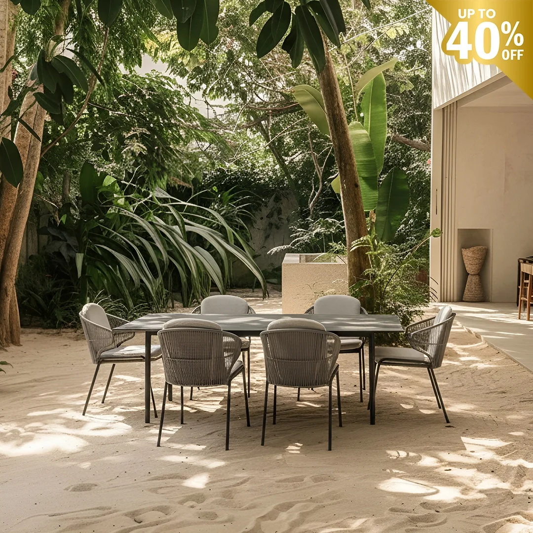 Outdoor dining area with six gray chairs around a black table on sandy ground, surrounded by lush green plants and trees, with a modern building in the background.