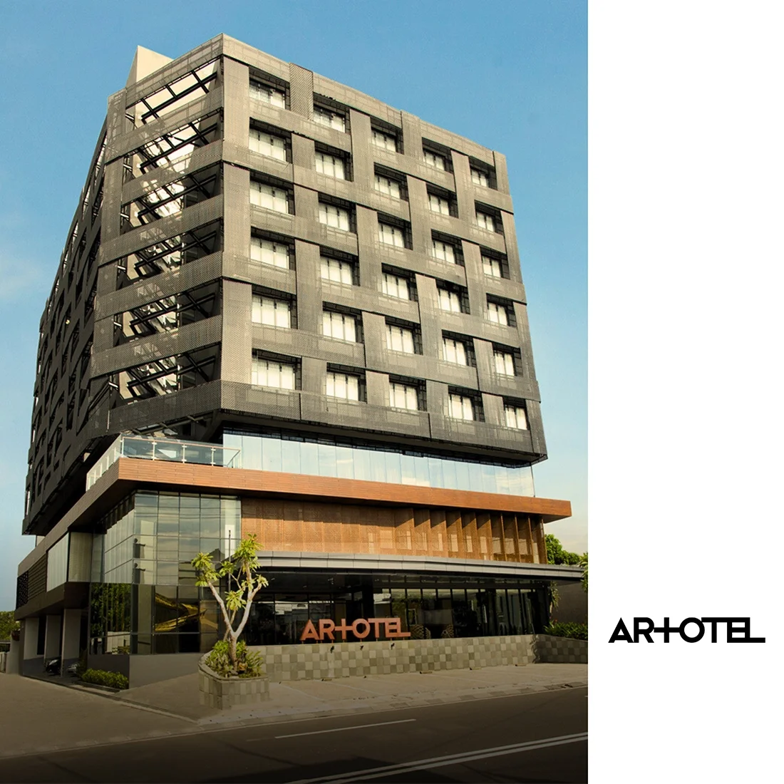 A modern multi-story hotel named AR!OTEL with a glass entrance, a wooden accent facade, and an exterior fire escape stairway on the left side of the building, set against a blue sky.