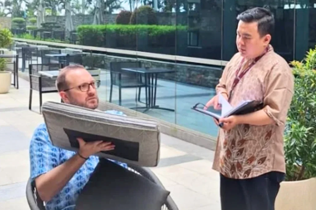 Man sitting in a wheelchair holding a cushion or pillow, talking to a woman standing nearby. The woman is holding a folder or notepad and appears to be explaining something. They are outdoors near a pool or water feature with glass barriers and outdoor furniture.