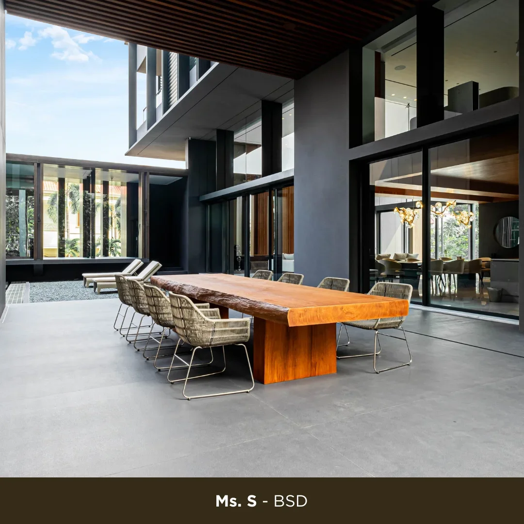 Modern outdoor patio with a wooden dining table and eight wicker chairs, adjacent to a contemporary building with large glass windows and sliding doors, overlooking a lush outdoor space.