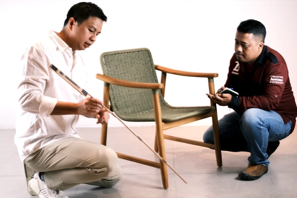 Two men, one kneeling and one sitting, examining a wooden chair with a woven seat. The man kneeling is holding a measuring stick, and the man sitting is taking notes.