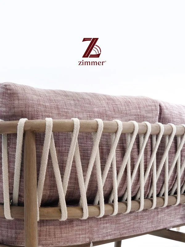 Close-up of a pink upholstered sofa with a wooden and rope decorative backrest, and a Zimmer logo on the wall above.