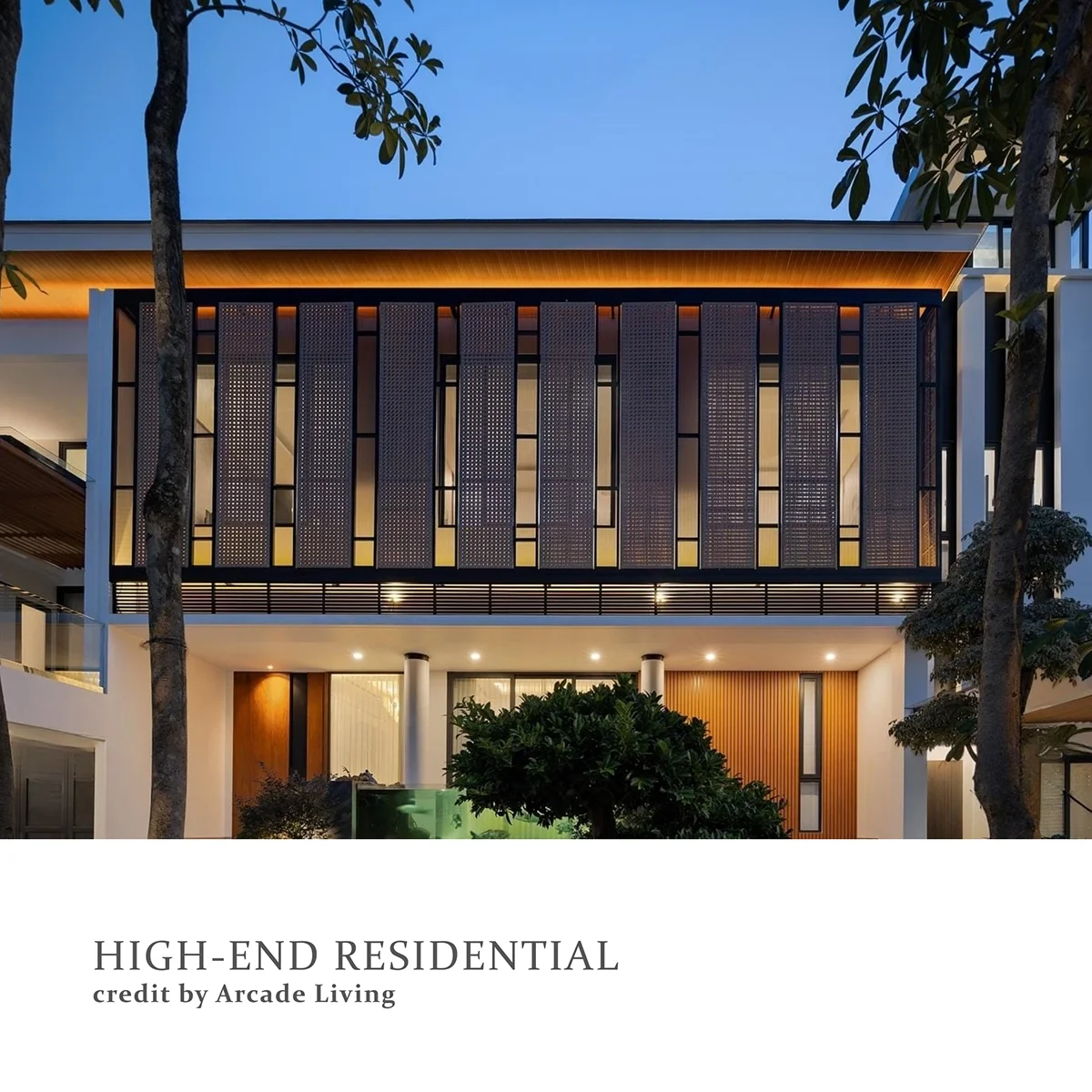 Exterior view of a modern high-end residential building at dusk with warm lighting, large windows, and decorative black metal facade panels. The building features trees and landscaping in the foreground.