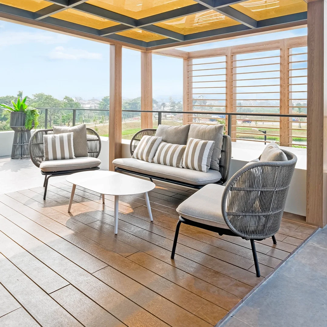 Outdoor patio with beige wicker lounge chairs, a beige cushioned sofa with striped pillows, a white coffee table, potted plant, and a wooden privacy screen with horizontal slats, overlooking a green landscape.