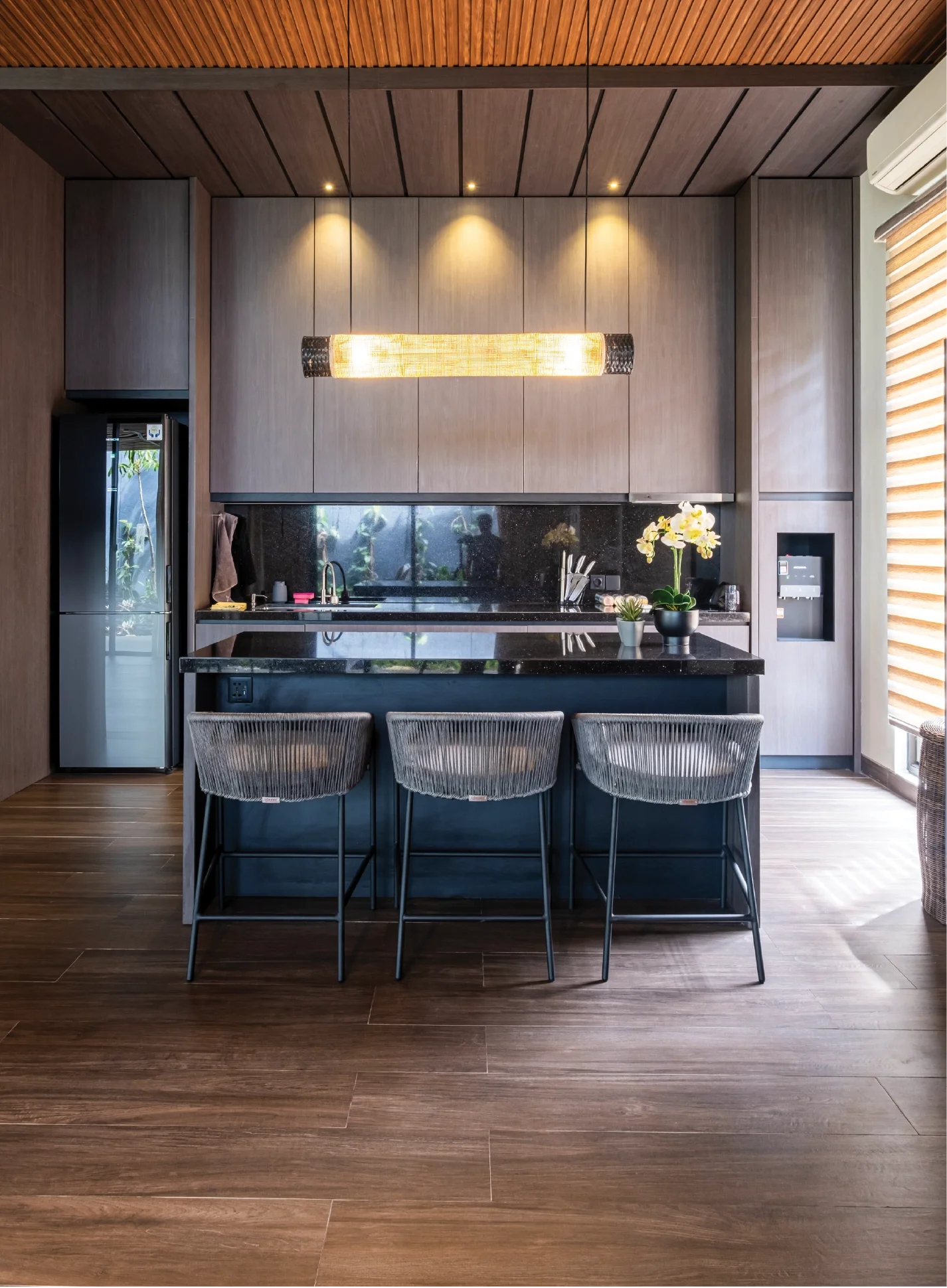 Modern kitchen with wooden cabinetry, black countertop, and three bar stools at the kitchen island. There is a refrigerator on the left, a sink, and decorative plants. Recessed lighting and a wooden ceiling add warmth to the space.