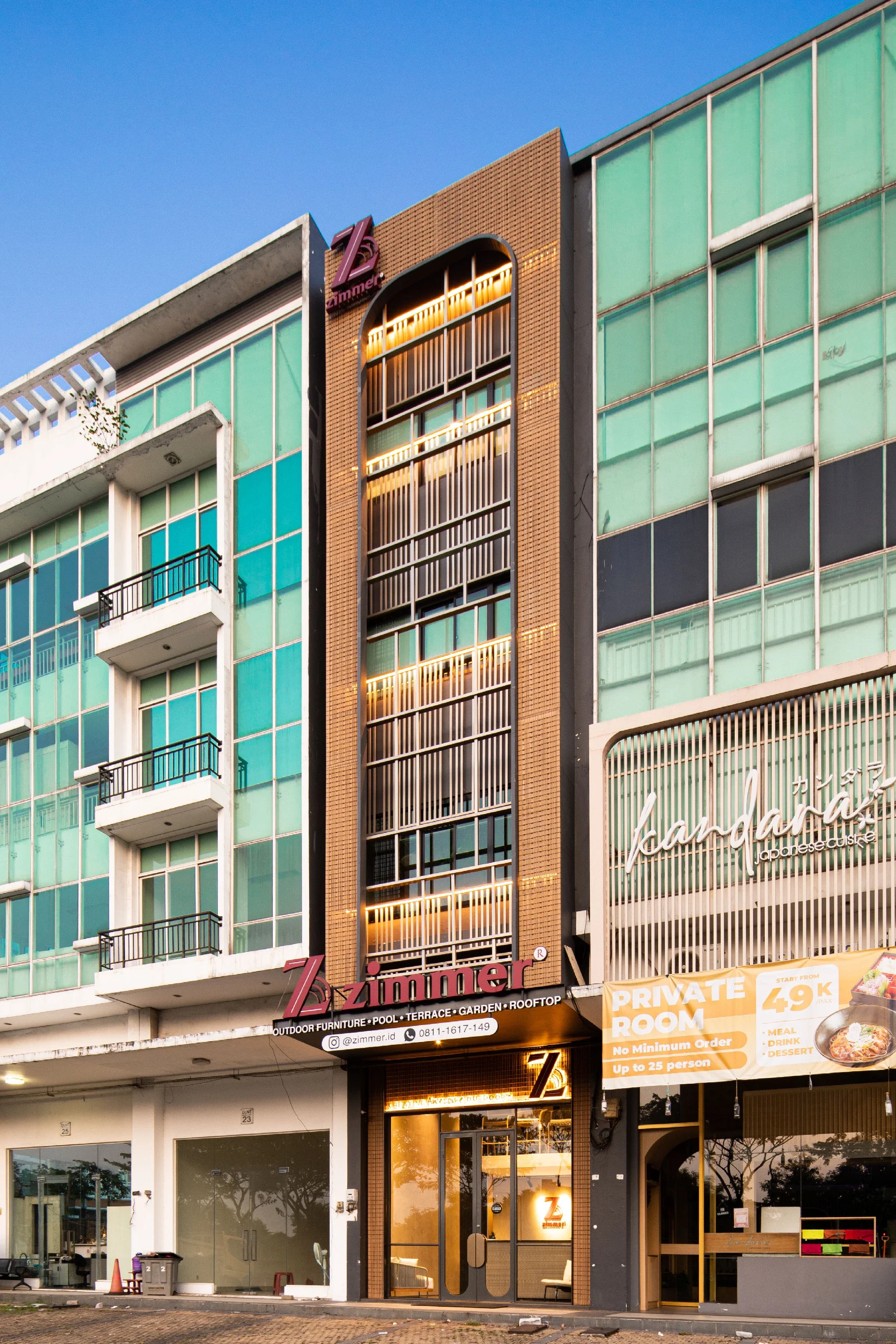 Multi-story building with a restaurant named Zimmer on the ground floor, featuring illuminated vertical signage and windows with warm lighting, situated beside other commercial buildings with glass facades.