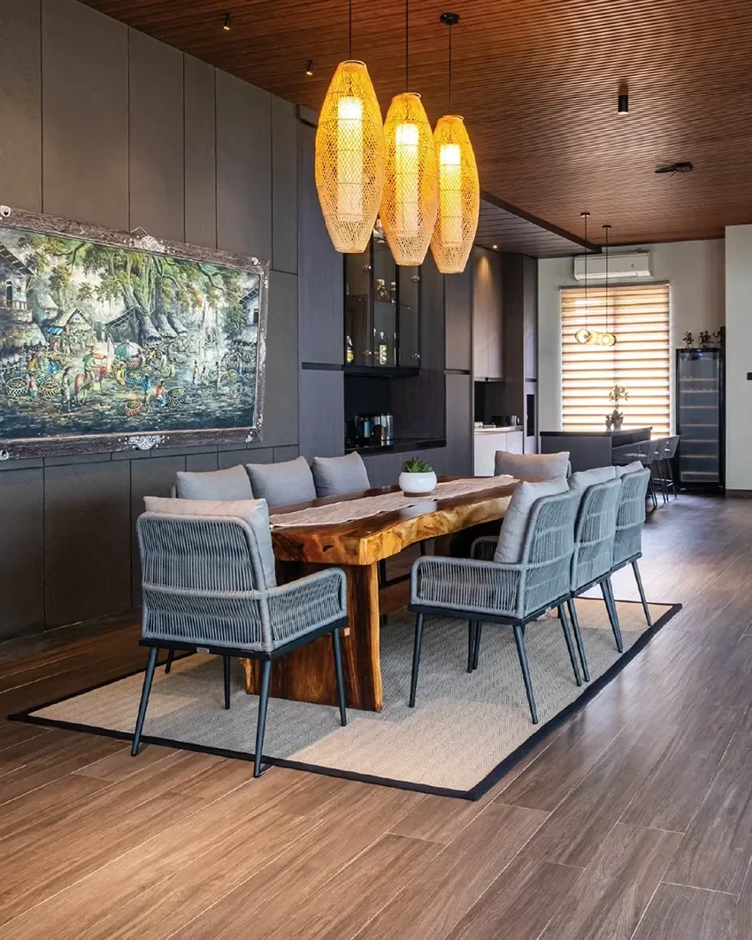 A dining room with a large wooden table, eight gray chairs, a beige rug, and a dark wall with a landscape painting. Hanging above are four wicker pendant lights. In the background, there is a window with blinds, a small counter, and a wine cooler.