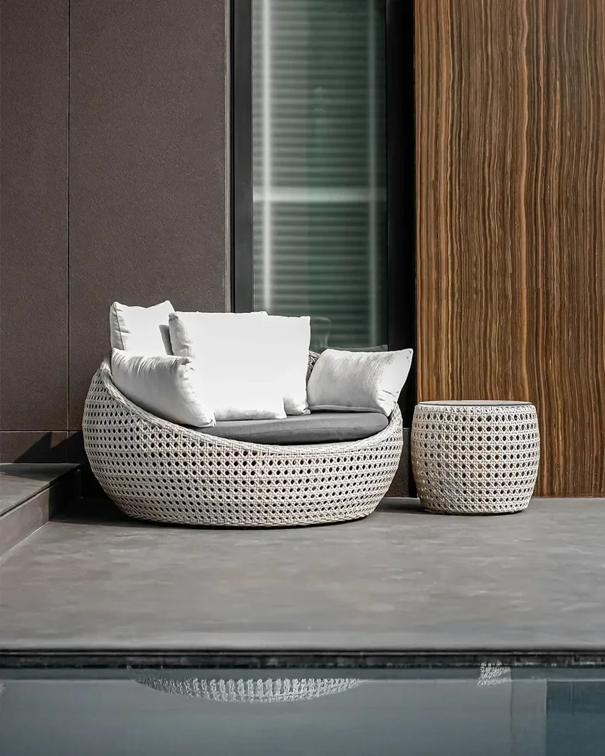 Modern outdoor seating with a round, woven area chair with gray cushions and matching woven side table, placed on a deck next to a swimming pool with a reflective surface, against a brown wall and glass window.