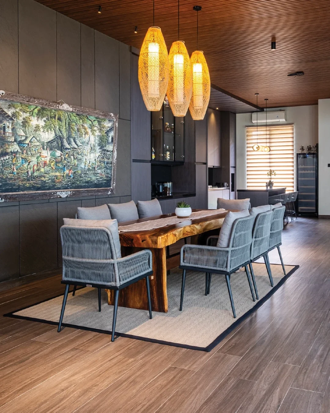 Modern dining room with a long wooden table surrounded by ten light gray upholstered chairs on a beige and black rug. Four woven hanging pendant lights illuminate the space. The dark wall on the left has a framed painting, with dark wood cabinetry and a window with wooden blinds in the background.
