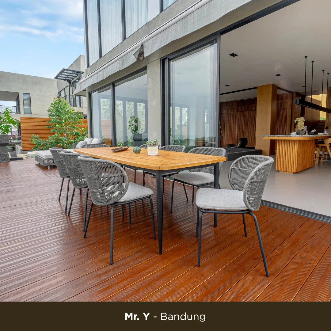 Outdoor patio with a wooden dining table and six gray chairs, next to a modern building with large glass windows, some greenery, and a kitchen area in the background.