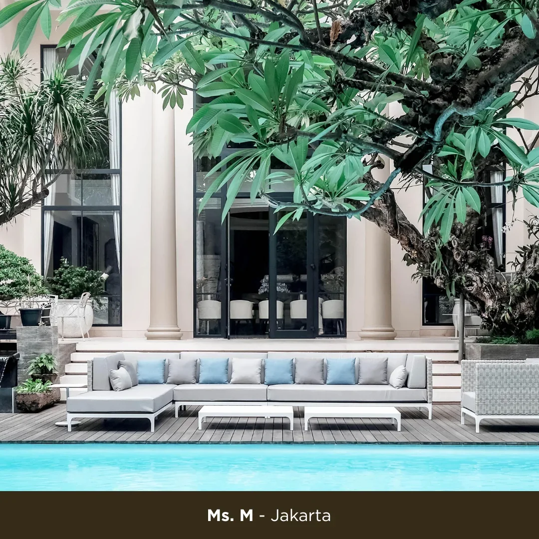 An outdoor lounge area with a modern gray sofa with blue and gray cushions, two small white coffee tables, and a pool in the foreground. There are tall windows and large greenery, including a large tree with broad leaves, surrounding the area.