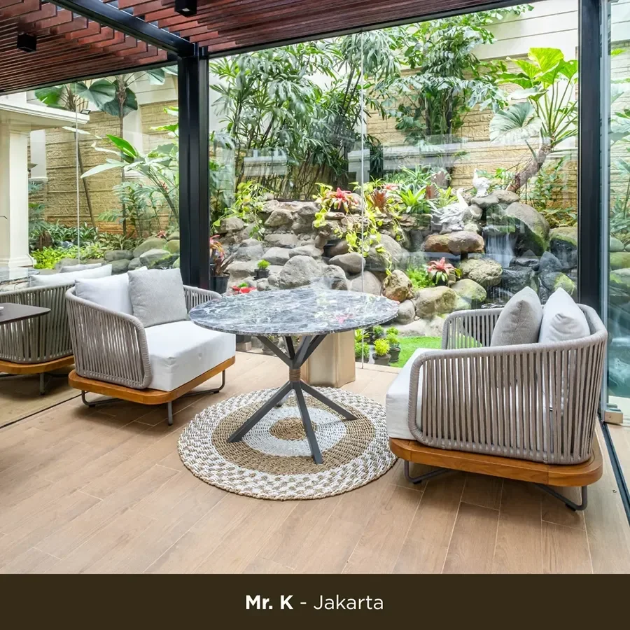 A cozy indoor seating area with two rattan chairs with white cushions, a round marble-topped table on a woven rug, and a large glass window revealing a lush garden with rocks, plants, and a small waterfall.