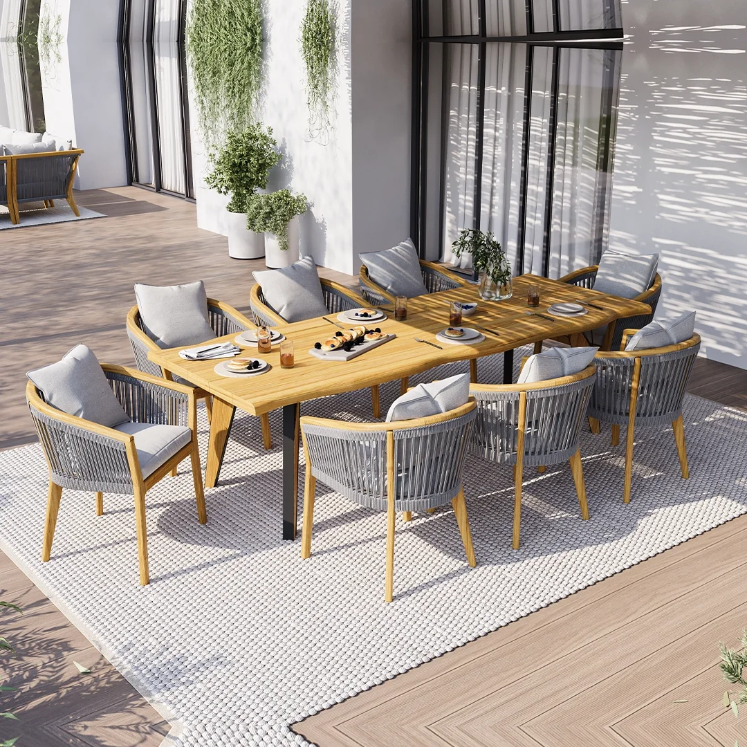 Dining room with a wooden table set with plates, glasses, and food, surrounded by chairs with cushions, on a white textured rug, with potted plants and large sliding glass doors allowing sunlight.