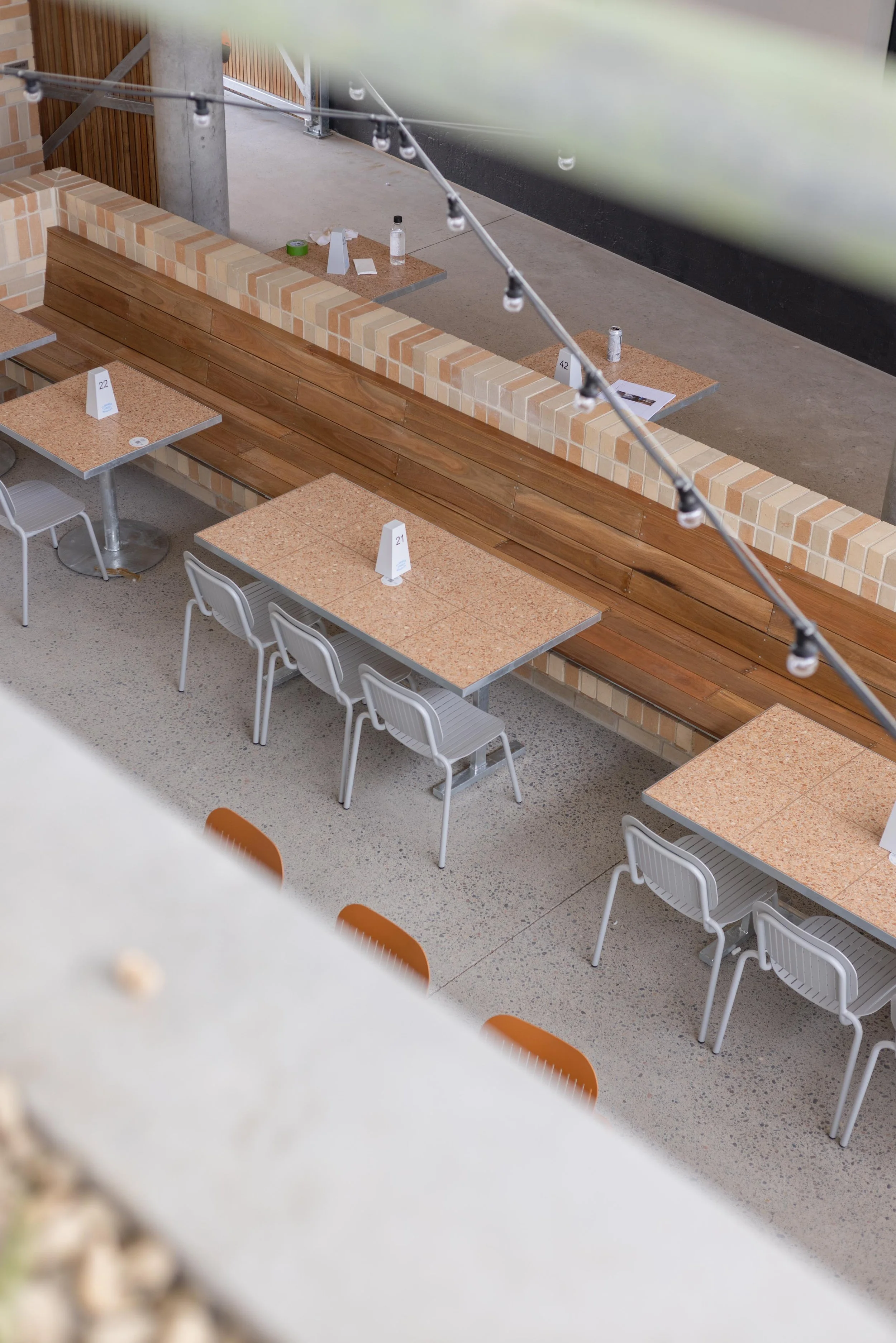 Indoor rooftop bar with a modern design, featuring a bar counter stocked with various bottles, tables with people socializing and drinking, large potted plants, and an outdoor terrace area with greenery and seating.