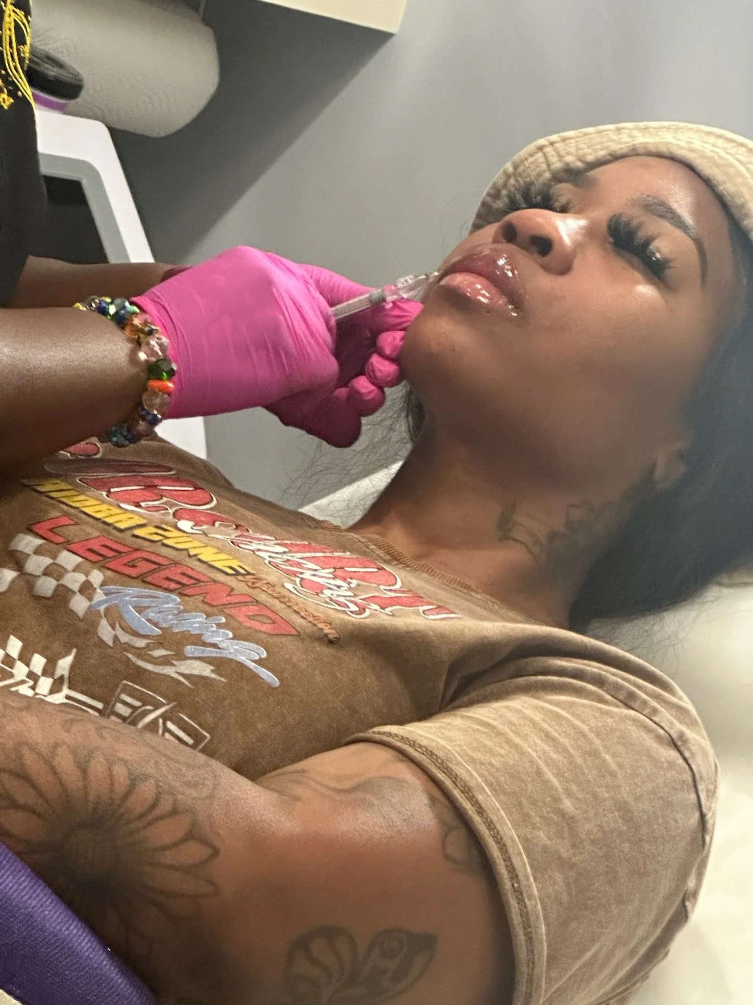 A woman with tattoos on her arm receiving a cosmetic injection in her chin from a healthcare professional wearing pink gloves. The woman has makeup, a beige hat, and a graphic brown T-shirt.