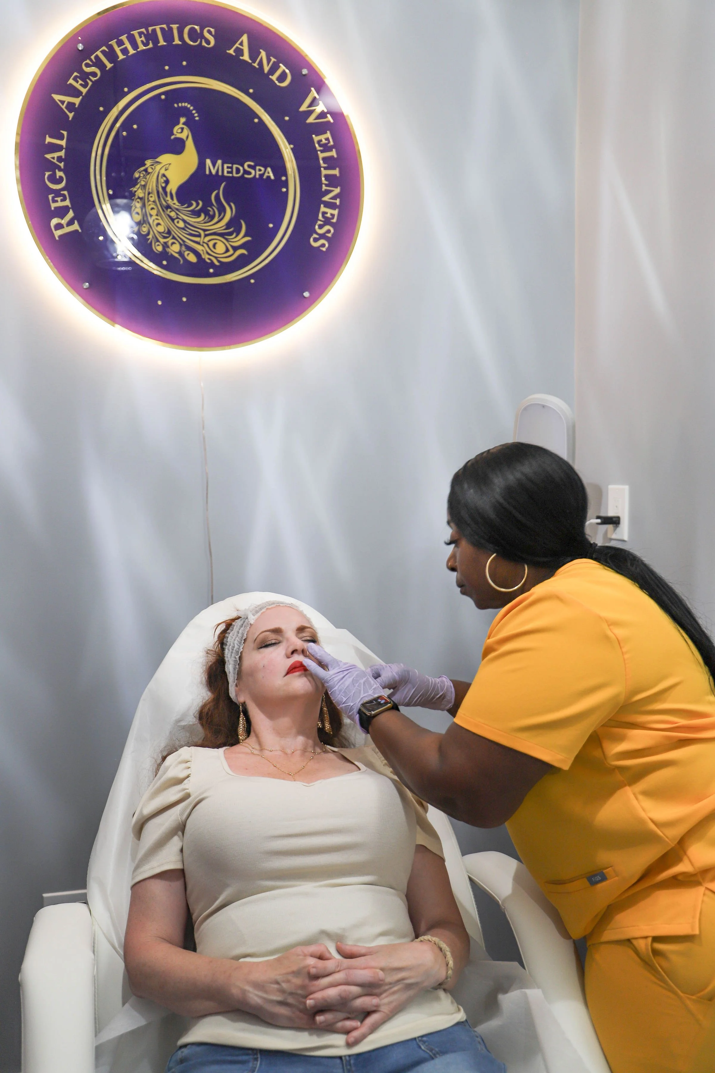 A woman receiving a cosmetic treatment on her face from a professional in a spa or clinic, with a sign for 'Regal Aesthetics and Wellness' in the background.