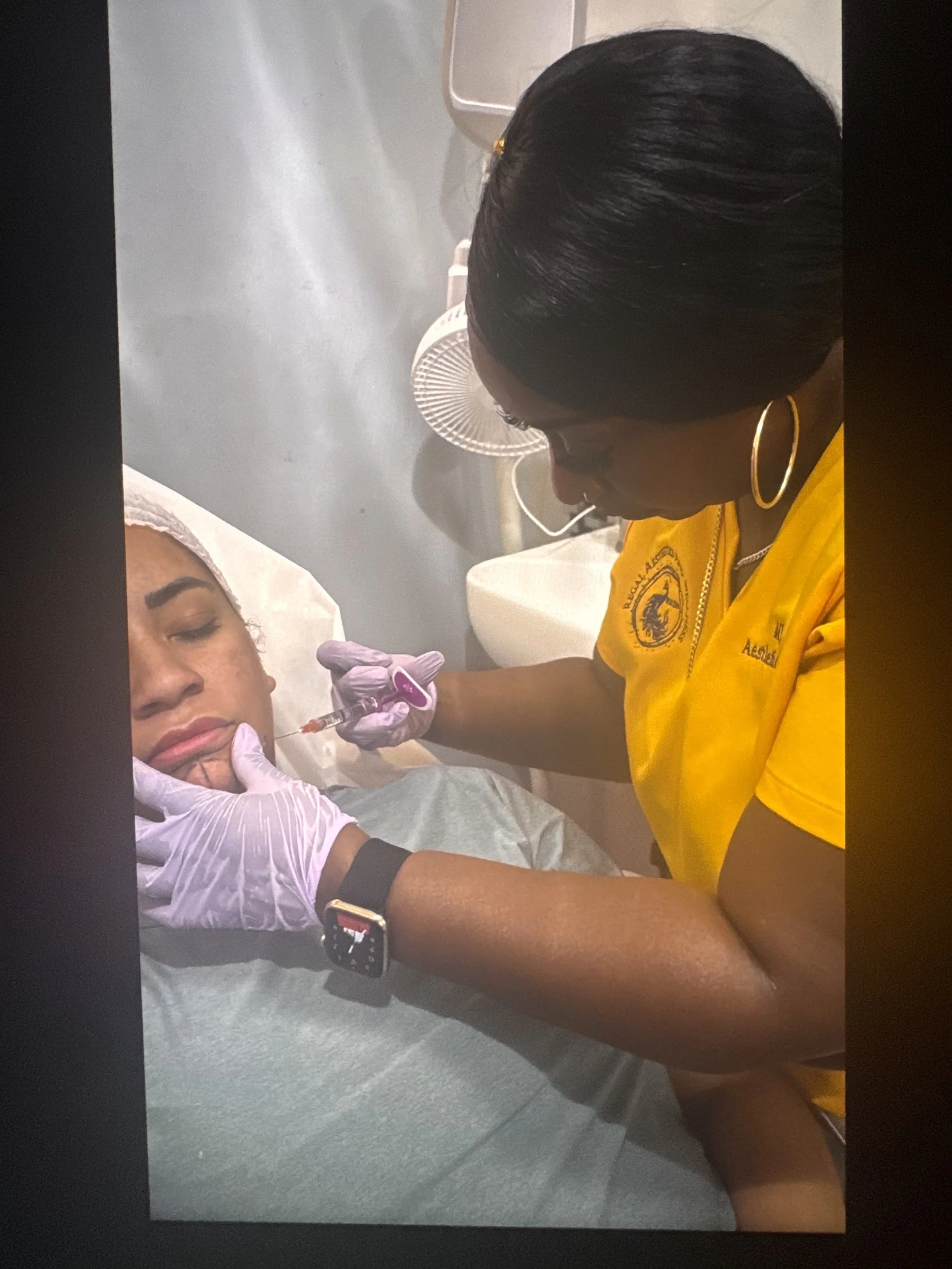 A woman receiving an injection in her chin while lying on a medical bed, another woman administering the shot, with a fan and medical equipment in the background.