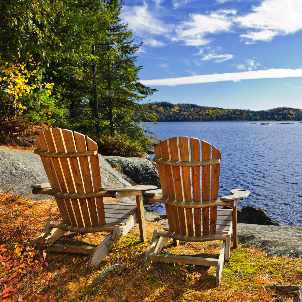 Two wooden Adirondack chairs face a large lake with a forested shoreline in the background and a partly cloudy blue sky overhead.