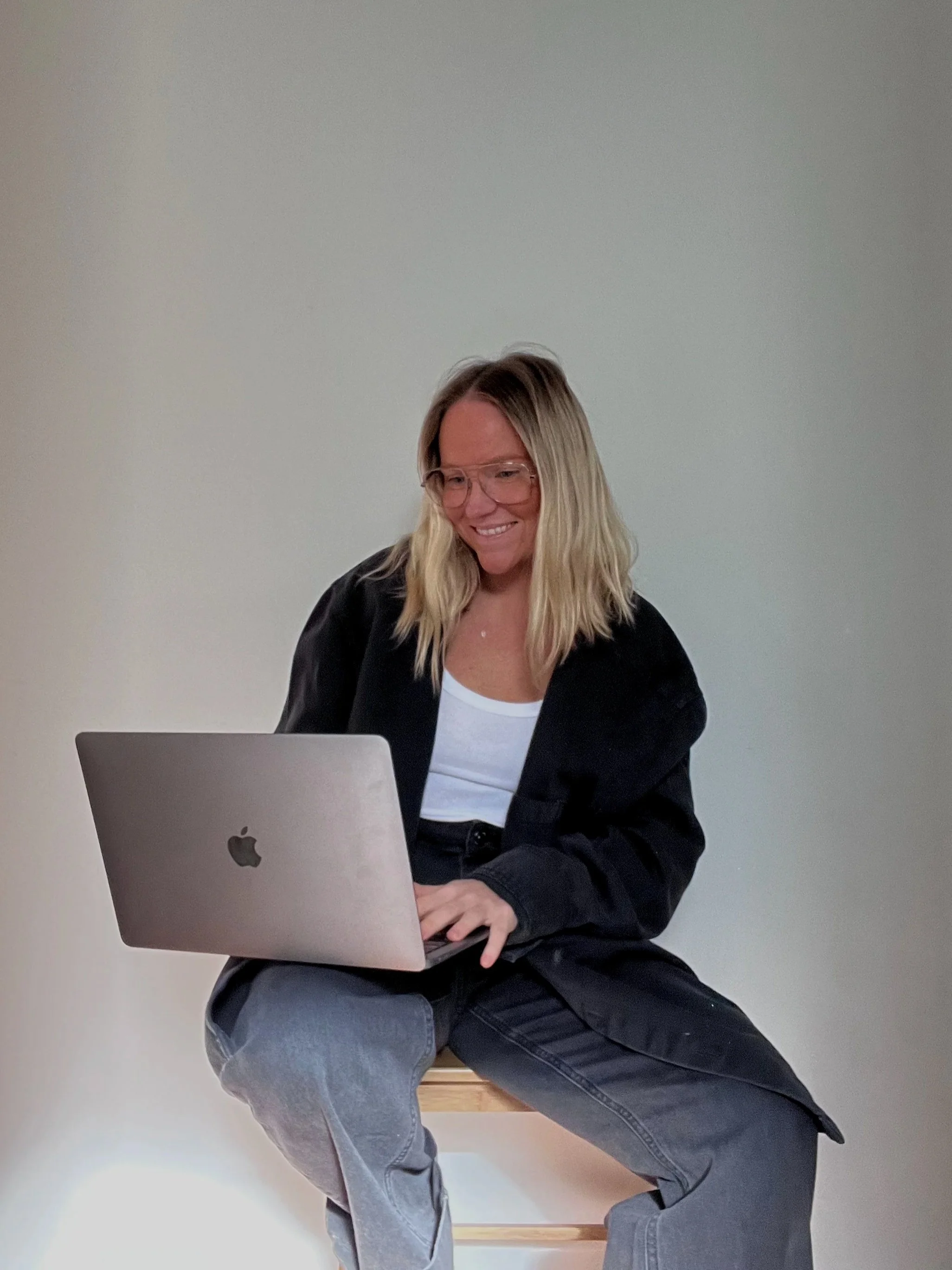 A woman with blonde hair, glasses, and a black jacket sitting on a bench using a silver MacBook laptop.