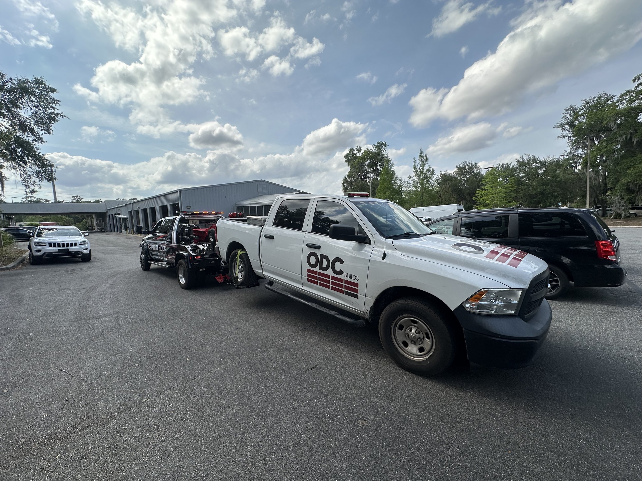 A white police pickup truck with 'ODC BUILDS' decal towing a black tow truck carrying a white police car with a 'POLICE' decal in a parking lot, with trees and a blue building in the background.