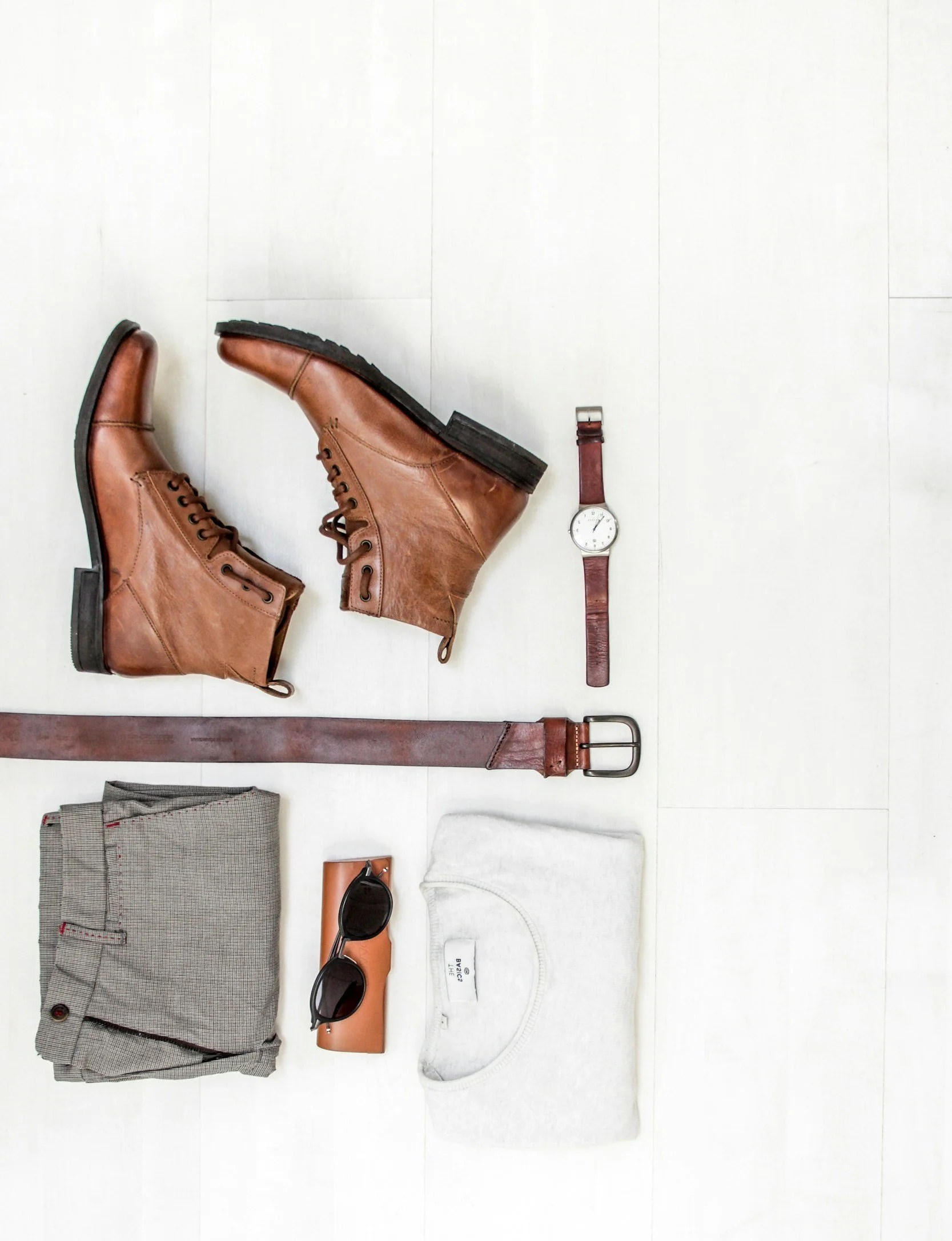 Brown leather boots, a brown leather belt, a watch with a brown strap, gray checked pants, black sunglasses on a tan case, a white T-shirt, and a pair of black sunglasses on a tan case arranged on a white background.