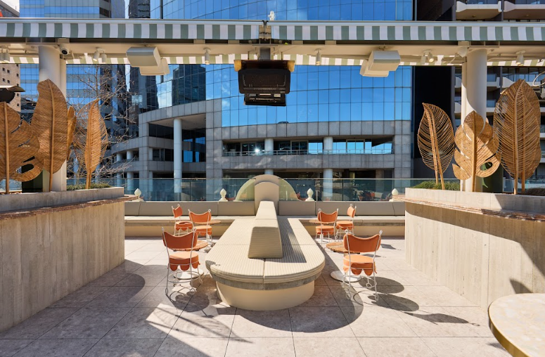 Outdoor rooftop seating area with orange chairs around a long curved built-in bench, large decorative brown and gold leaf-shaped planters, and modern glass buildings in the background. The Bristol Hotel in Sydney, Charitabl. Launch Party Promo.
