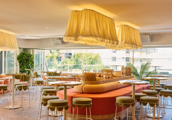 Bright, modern interior of a cafe or lounge with round tables, cushioned seating, large fabric light fixtures, and large windows with a view of trees and buildings outside. The Bristol Hotel in Sydney, Charitabl. Launch Party Promo.