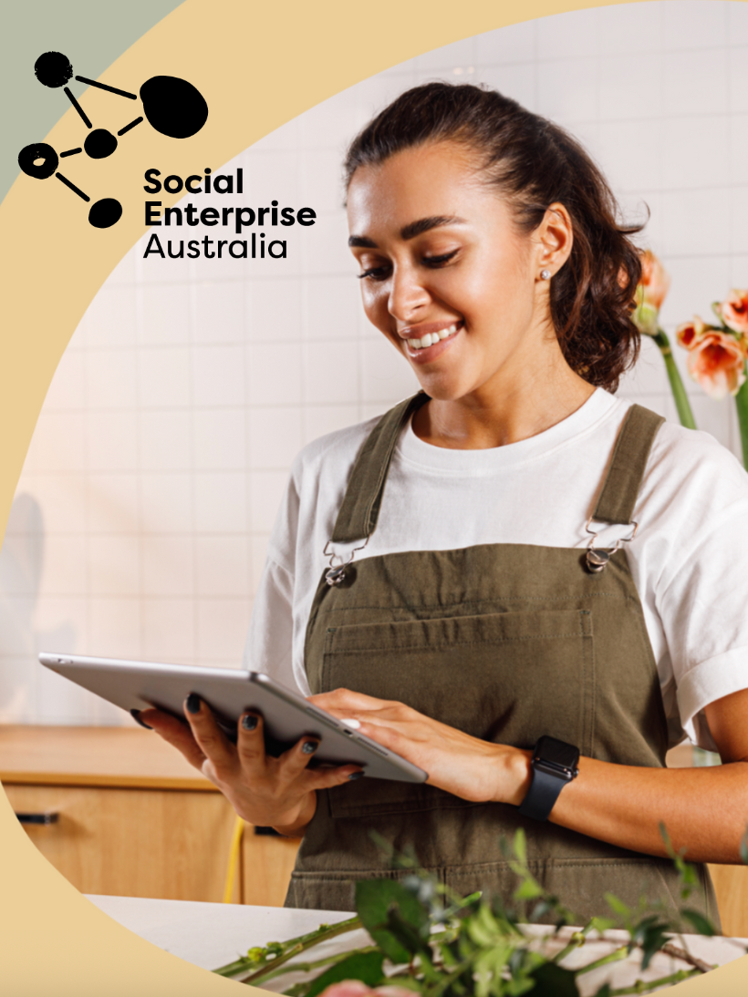 A woman in a white T-shirt and green apron smiling while using a tablet in a kitchen with flowers in the background. Social Enterprise Australia.
