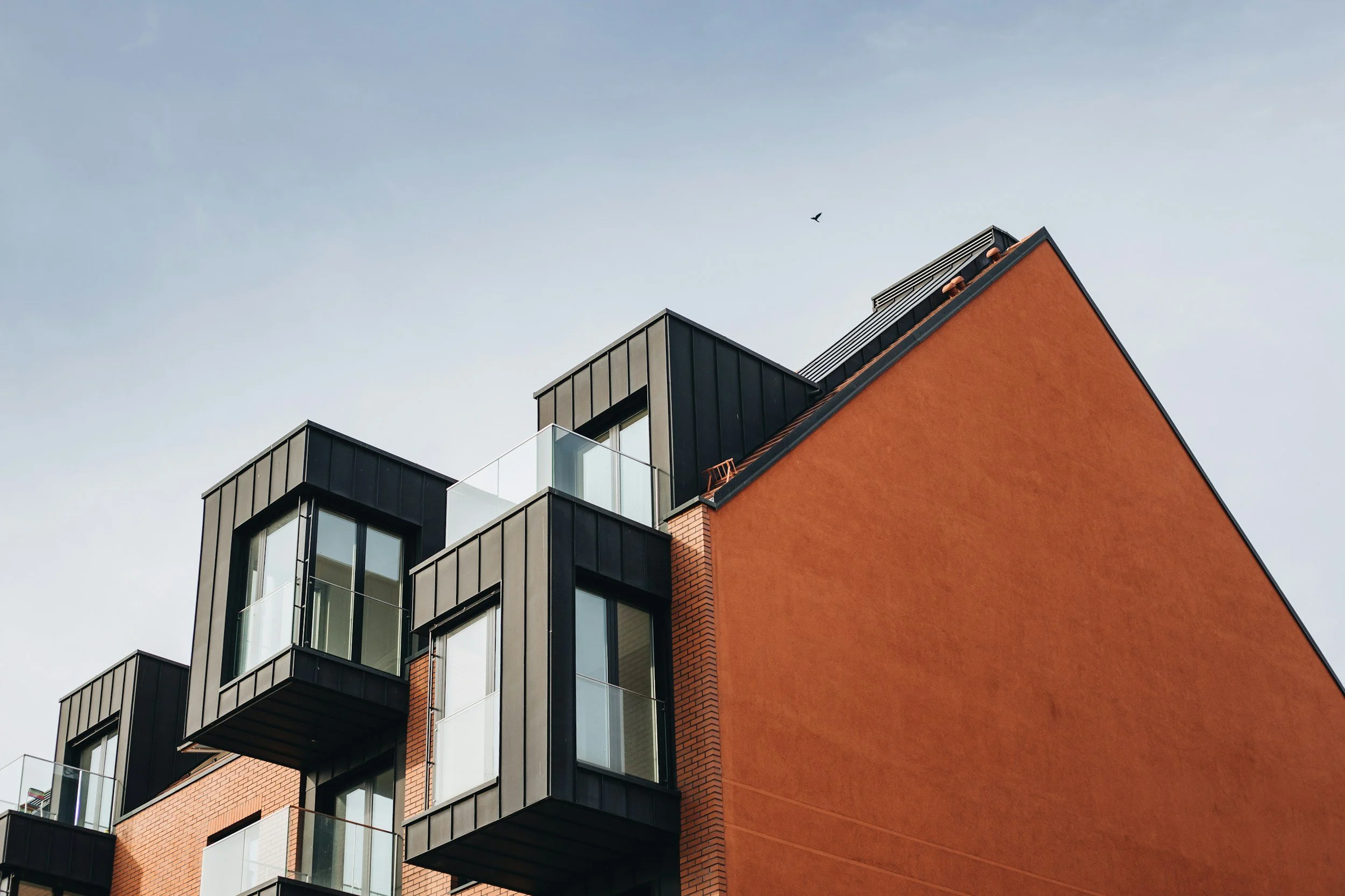 Modern apartment building with black metal and brick exterior, featuring protruding glass balcony sections and a large sloped orange wall, against a pale sky with a bird flying.