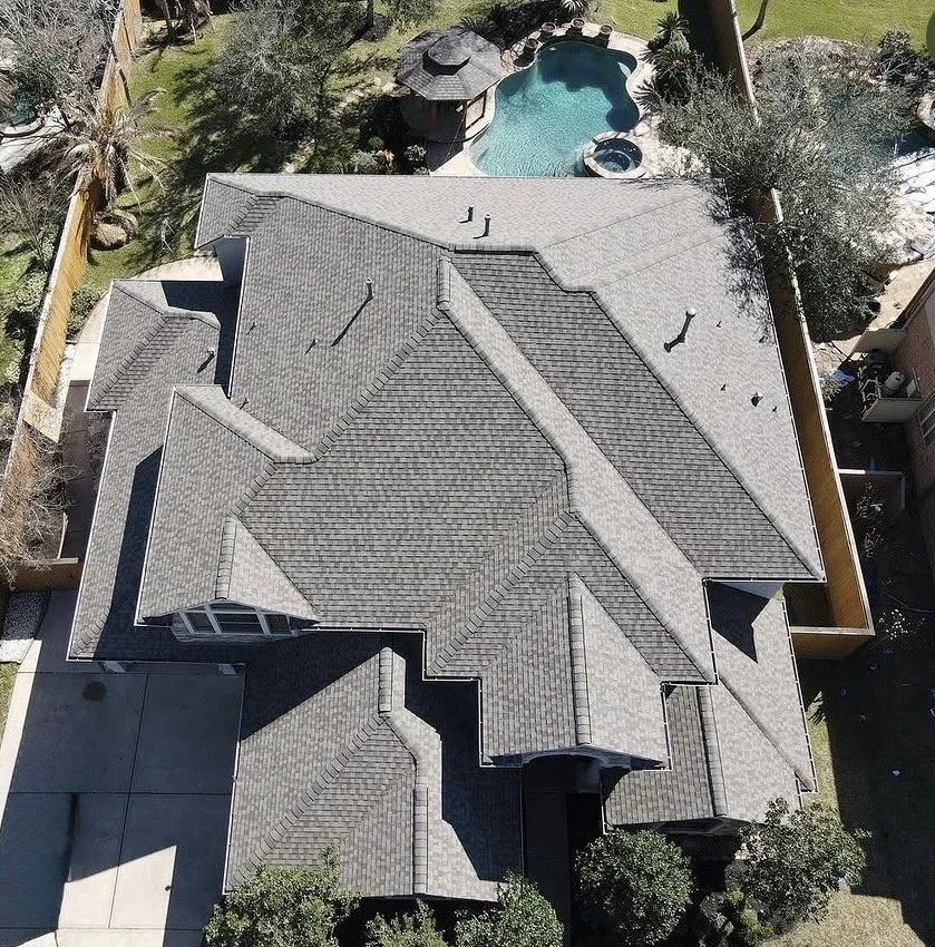 Aerial view of a house with a complex gray shingle roof, backyard, and a swimming pool surrounded by trees.