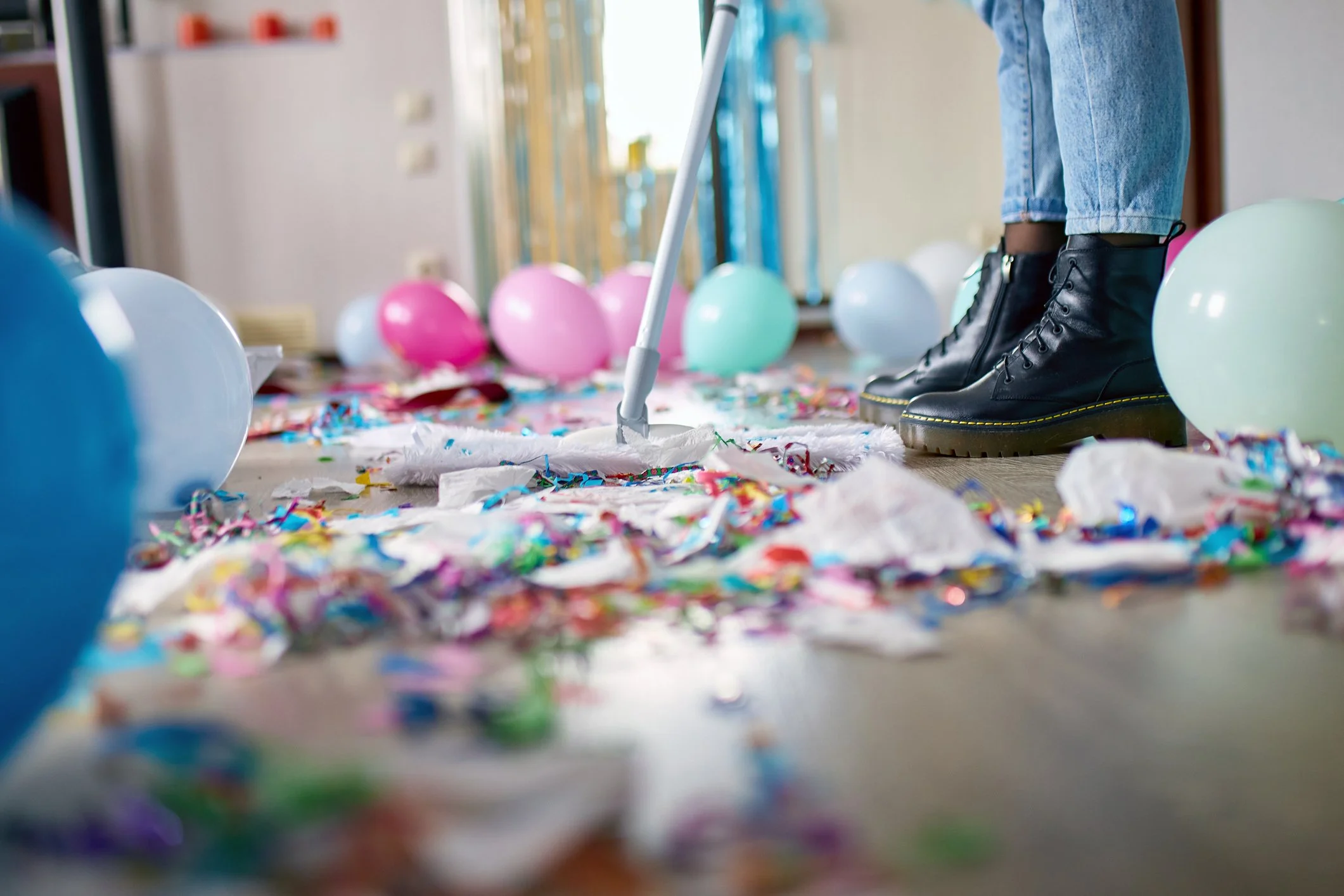 Person cleaning up colorful party confetti, balloons, and trash from the floor with a mop.