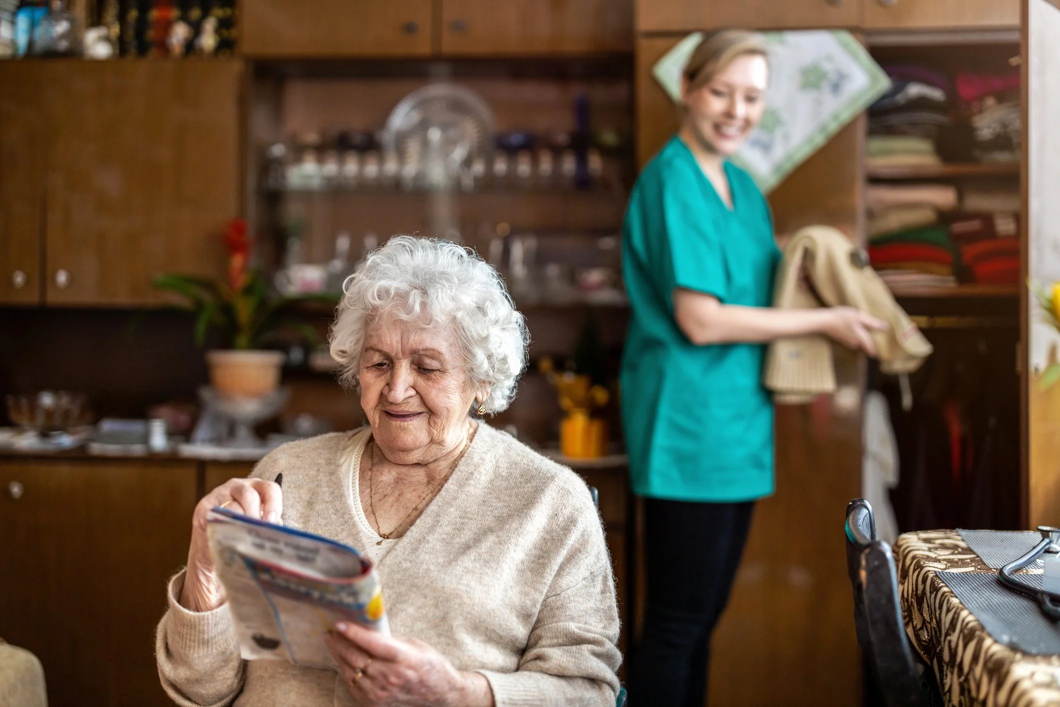 An elderly woman with curly white hair reads a magazine at a dining table, while a caregiver in green scrubs holds a folded towel and a pillow in the background, in a cozy room with wooden cabinets and shelves.
